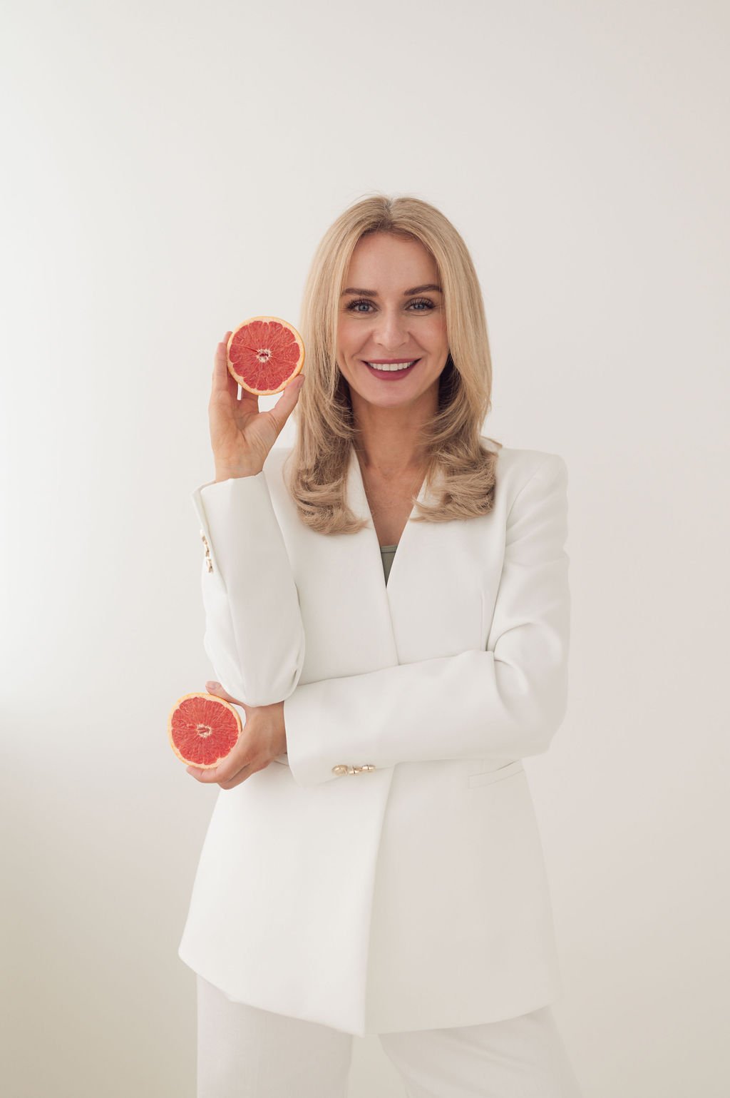 A woman with blonde hair wearing a white blazer holds a halved grapefruit in each hand, smiling at the camera against a plain white background.