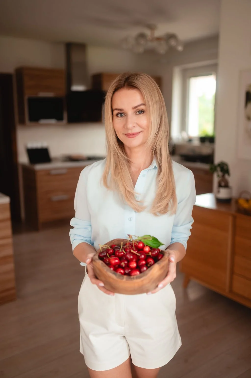 A woman with blonde hair, wearing a light blue shirt and white shorts, is smiling and holding a wooden bowl filled with fresh red cherries in a modern kitchen.