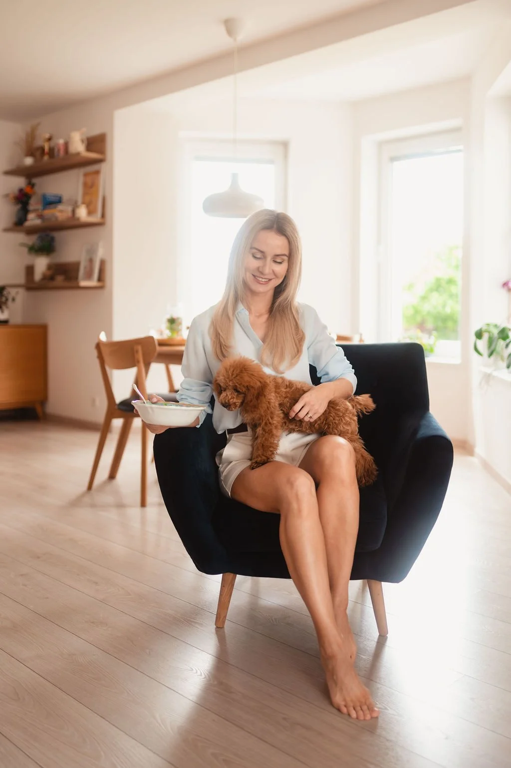 A woman sitting in a black armchair in a bright living room, holding a small brown poodle on her lap while smiling and eating from a bowl.