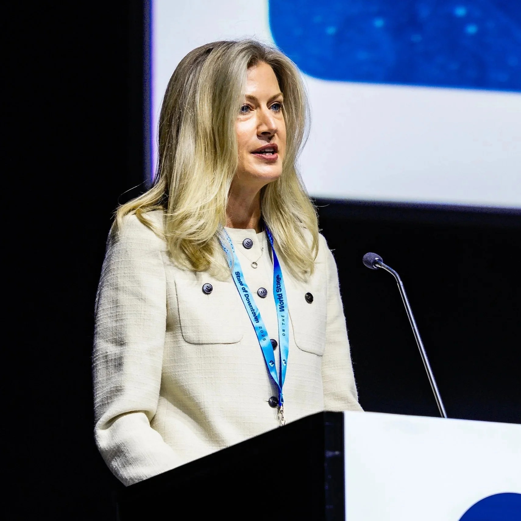 A woman with blonde hair, wearing a beige blazer, speaking at a podium with a microphone. She's wearing a blue lanyard labeled "World on the Move" and appears to be giving a speech or presentation at a conference or event.
