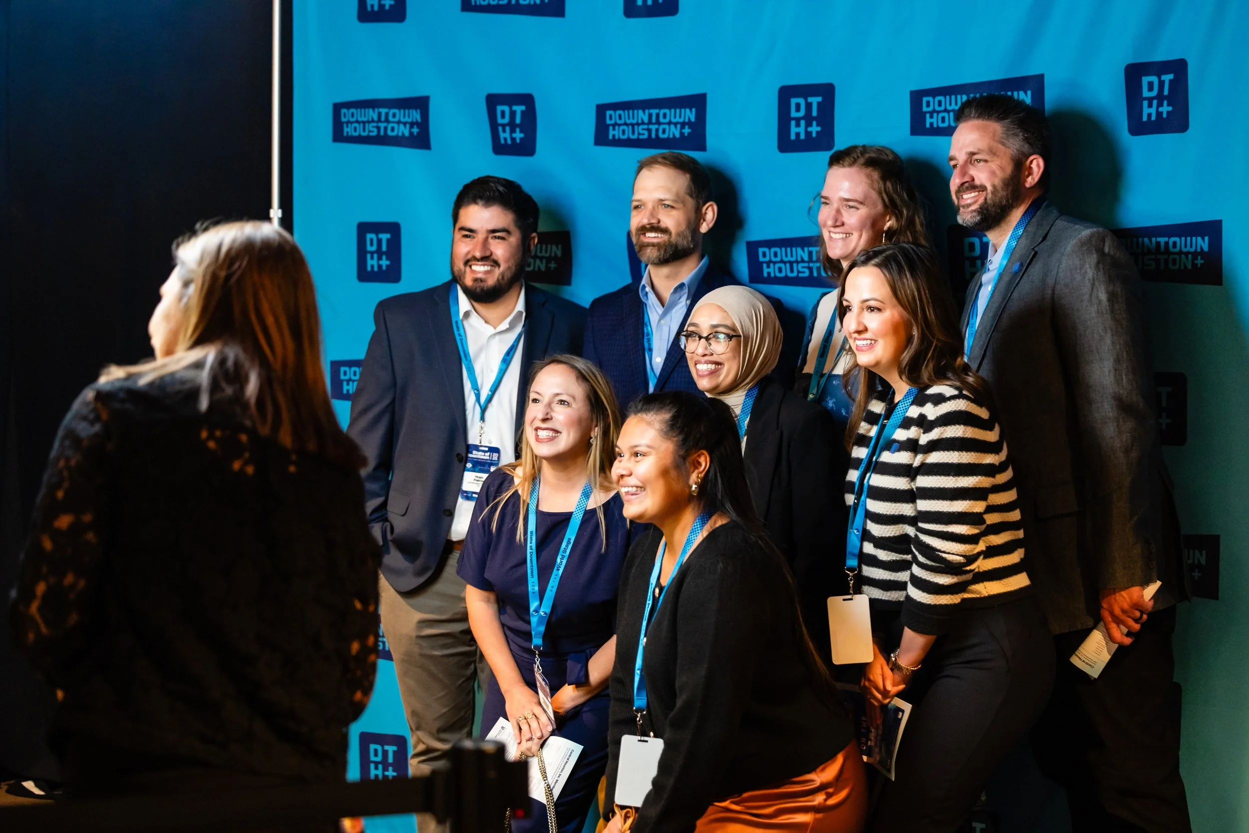 Group of diverse professionals smiling and posing for a photo at a conference with a blue backdrop displaying 'Downtown Houston+' logos.