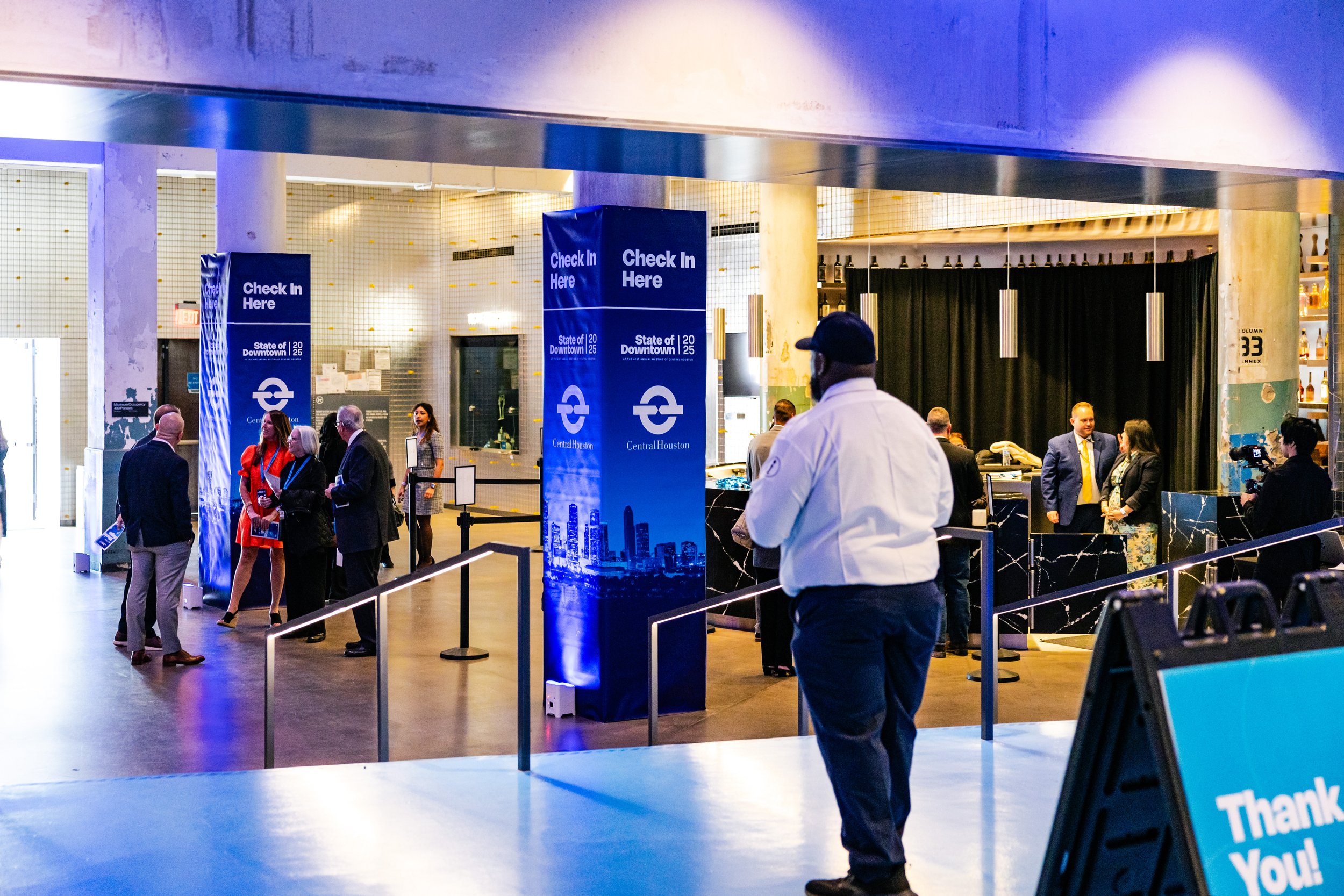 People at a check-in counter at an airport or train station, with a large blue sign that says 'Check In Here' and displays 'State of Downtown 2025.' The area appears busy with several individuals standing and talking, some with lanyards and badges. There are black curtains, shelves with bottles, and a person in a white shirt and cap visible in the foreground.