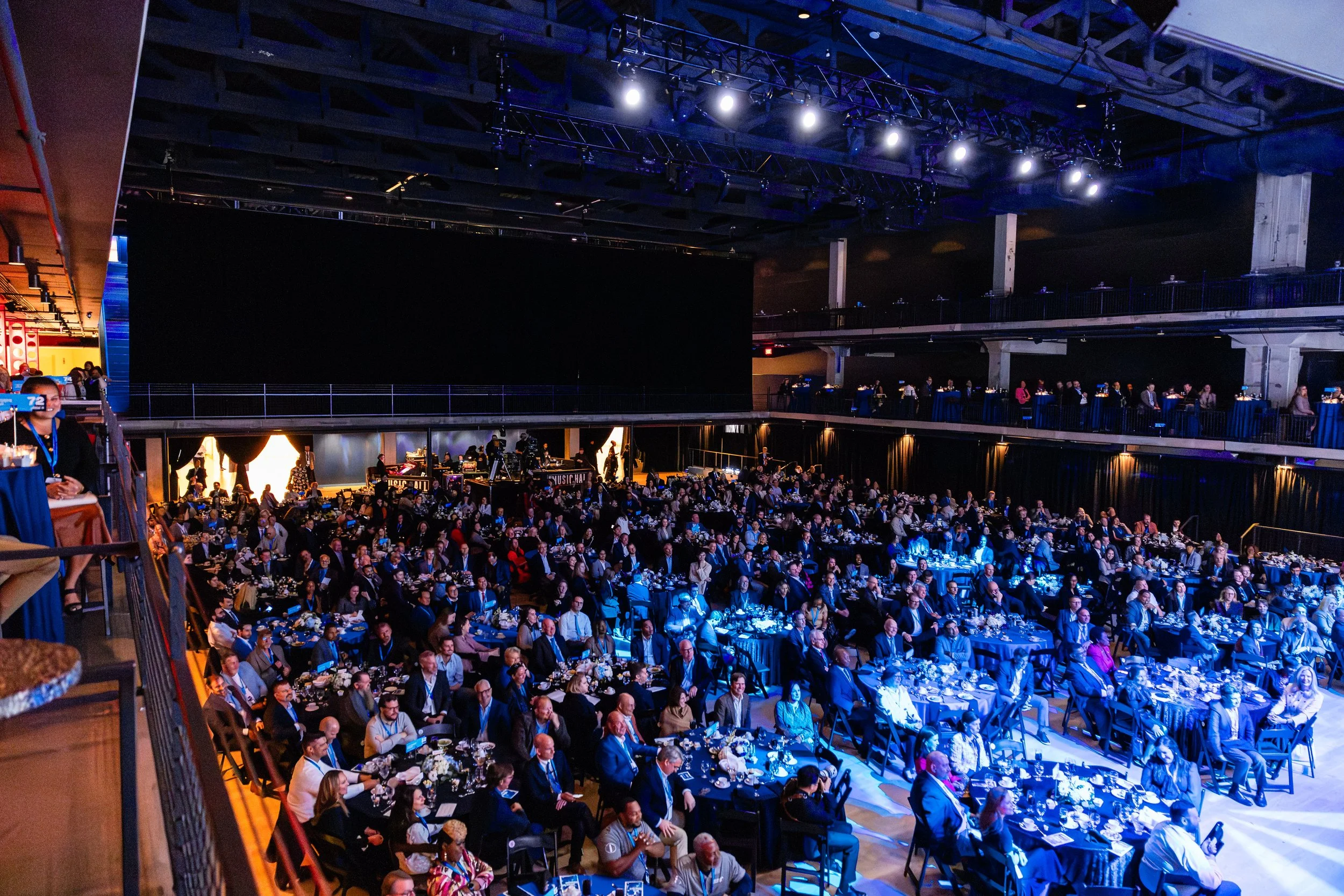 An indoor conference or banquet hall filled with attendees seated at round tables, with some on a balcony level, and a large black screen or stage at the front, illuminated by overhead lights.
