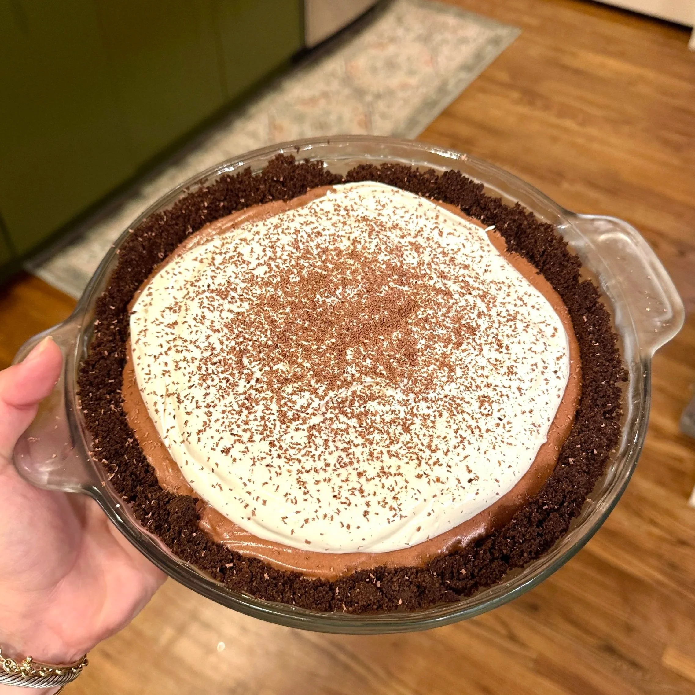 Close-up of a chocolate dessert in a glass dish with whipped cream and chocolate shavings, surrounded by crushed cookies on the edges.