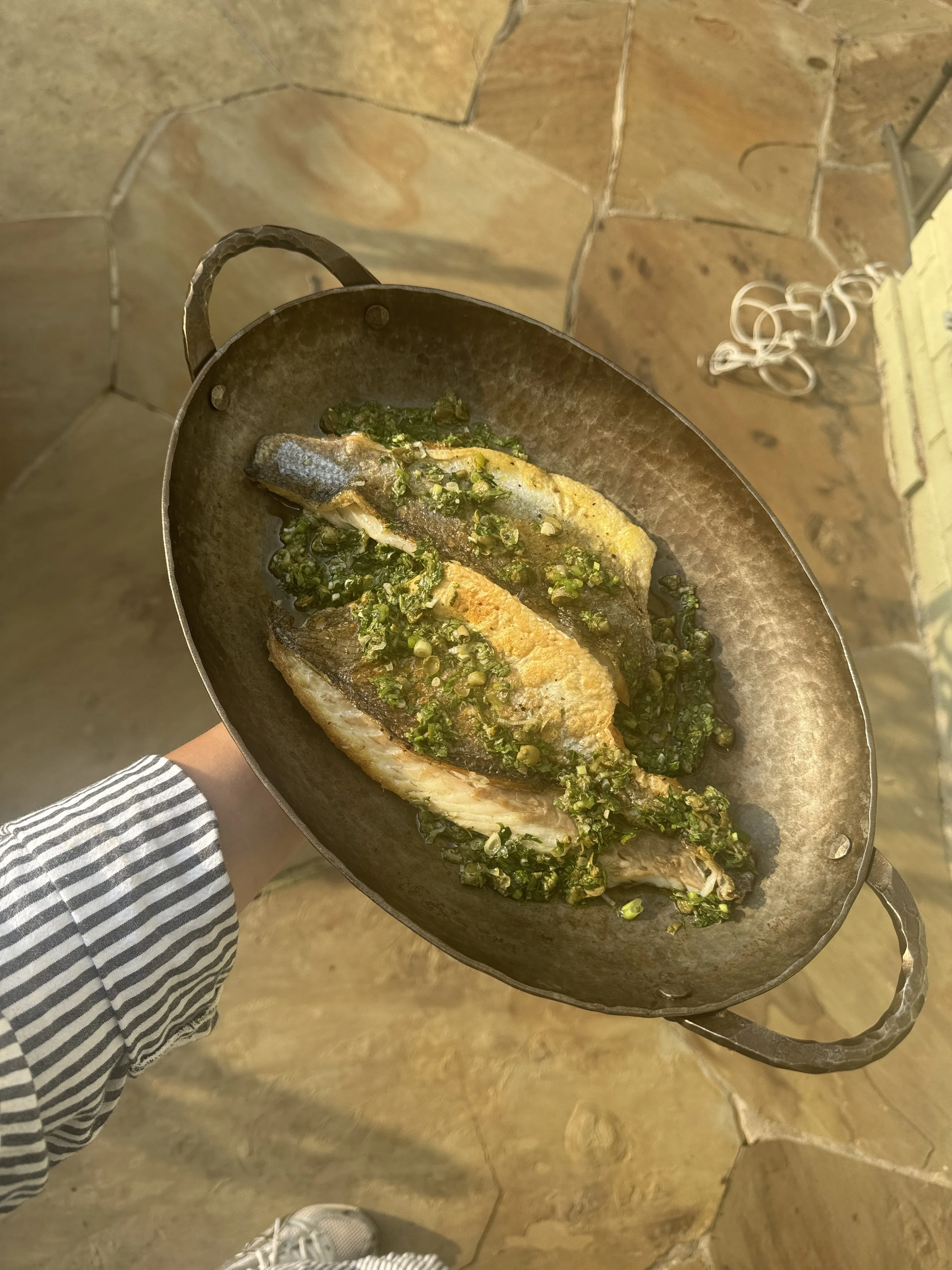 Person holding a pan with cooked fish topped with green herbs on a rustic stone floor.
