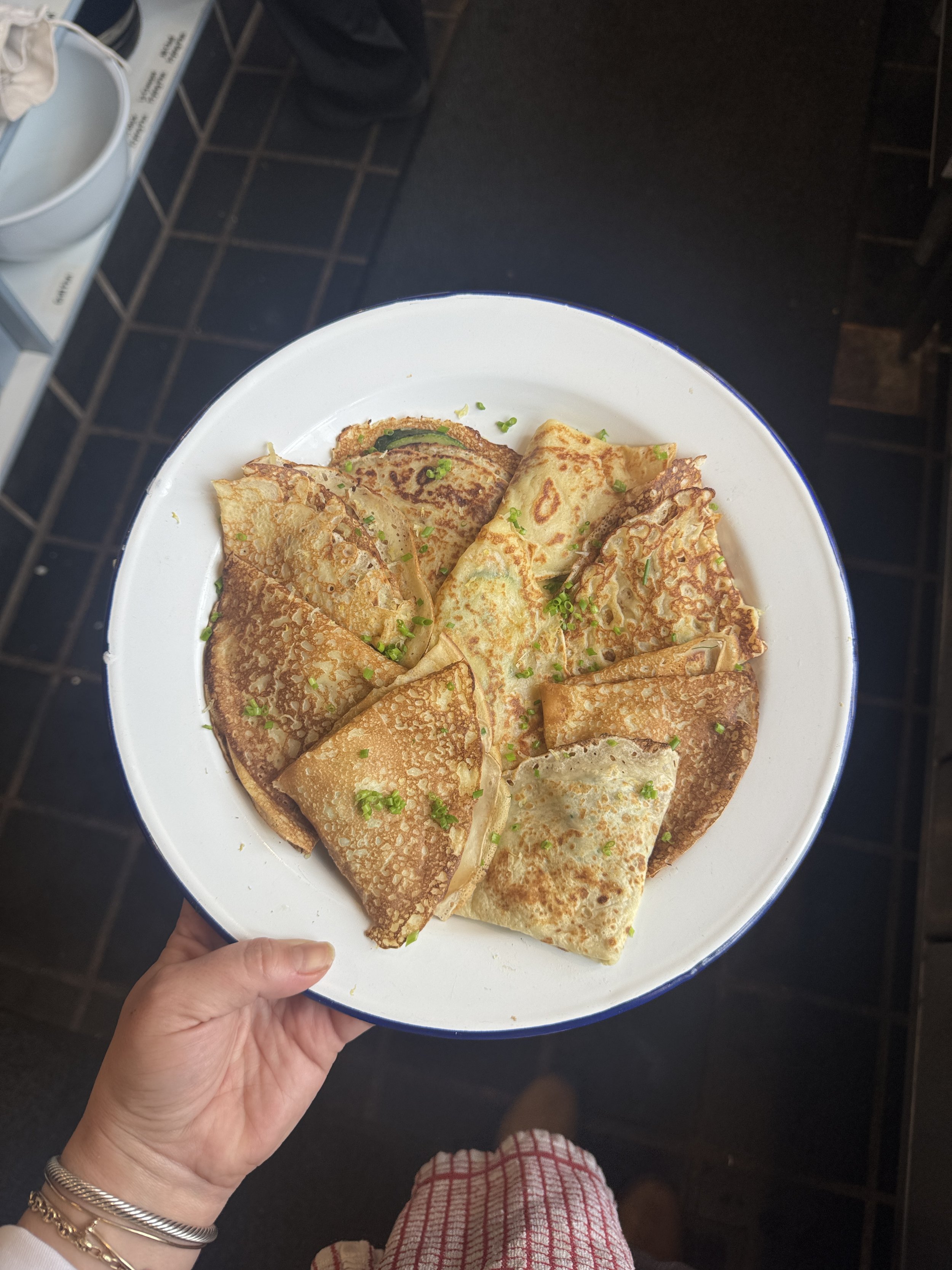 A hand holding a white plate with several folded crepes garnished with chopped green onions. The person is wearing multiple gold bracelets on their wrist, and the photo is taken in what appears to be a casual restaurant or kitchen setting with black tiled floors.