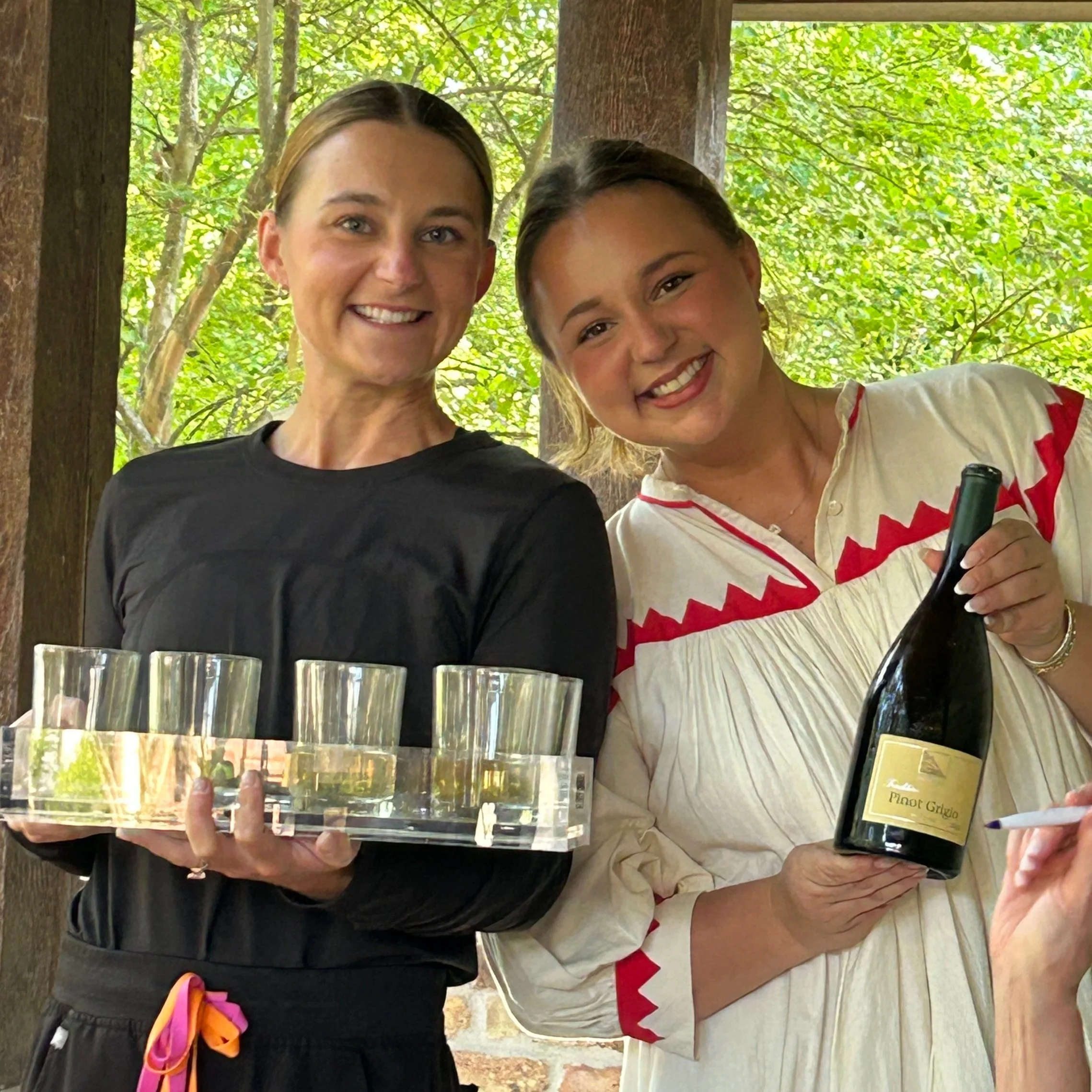 Two women smiling and posing for a photo in an outdoor setting with green tree foliage in the background. One woman is holding a large sheet of paper or a framed photo, and the other has her arm around her. There are glasses, a wine bottle, and a glass on the table in front of them.