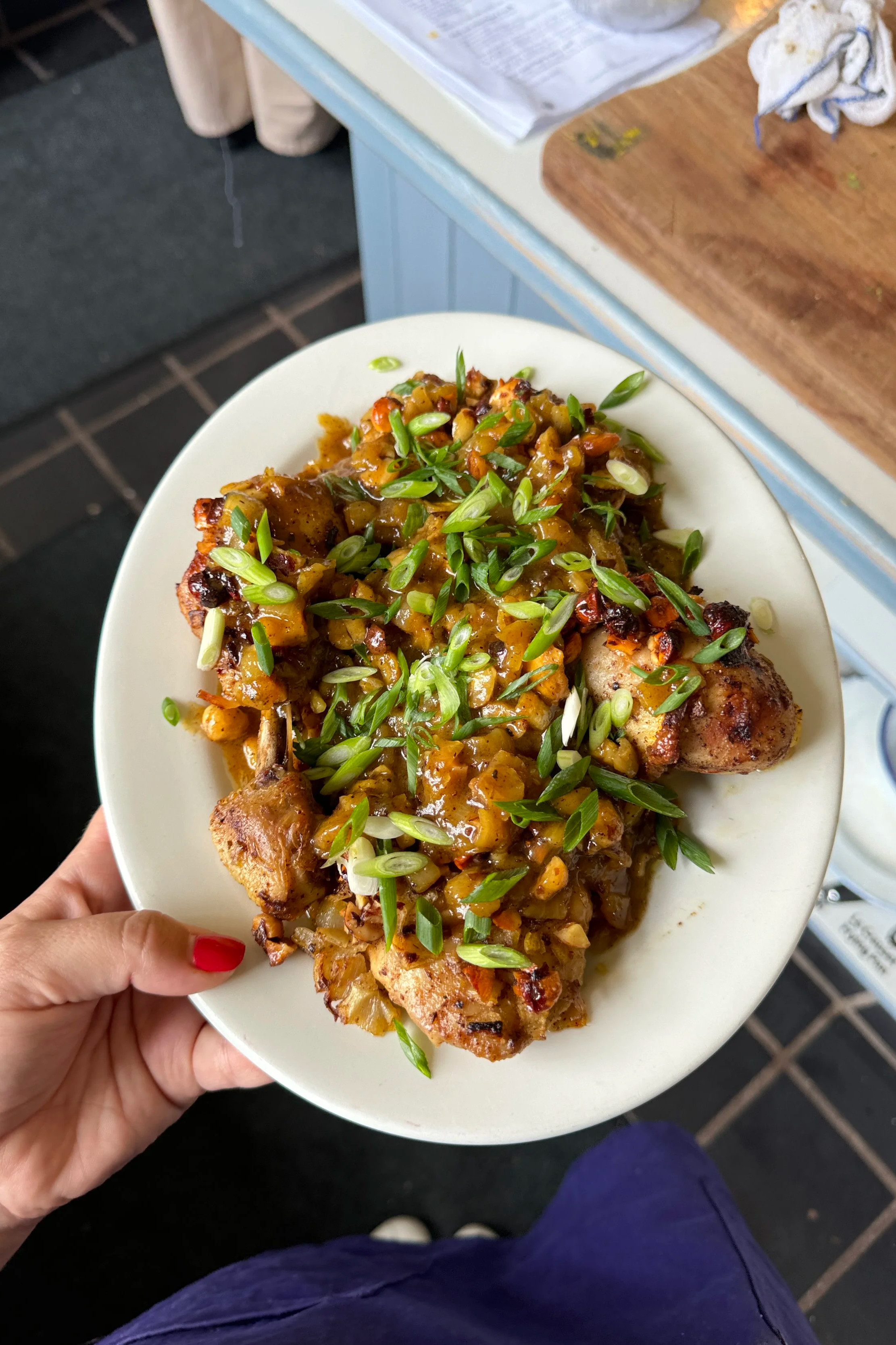 Close-up of a white plate with grilled chicken topped with a chunky vegetable sauce and garnished with sliced green onions, held by a hand with red-painted nails.