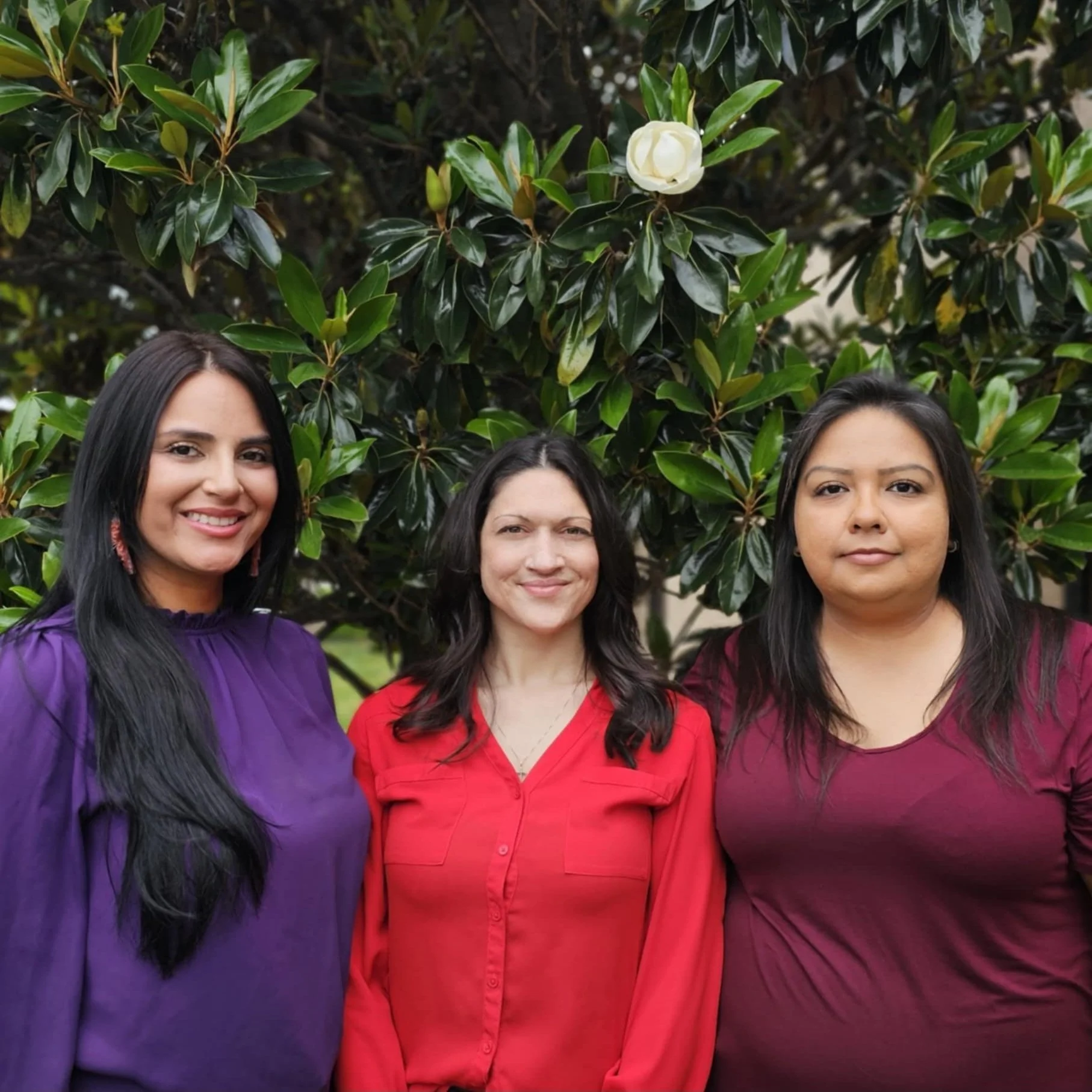 Three women standing outdoors in front of a leafy plant with a white flower.