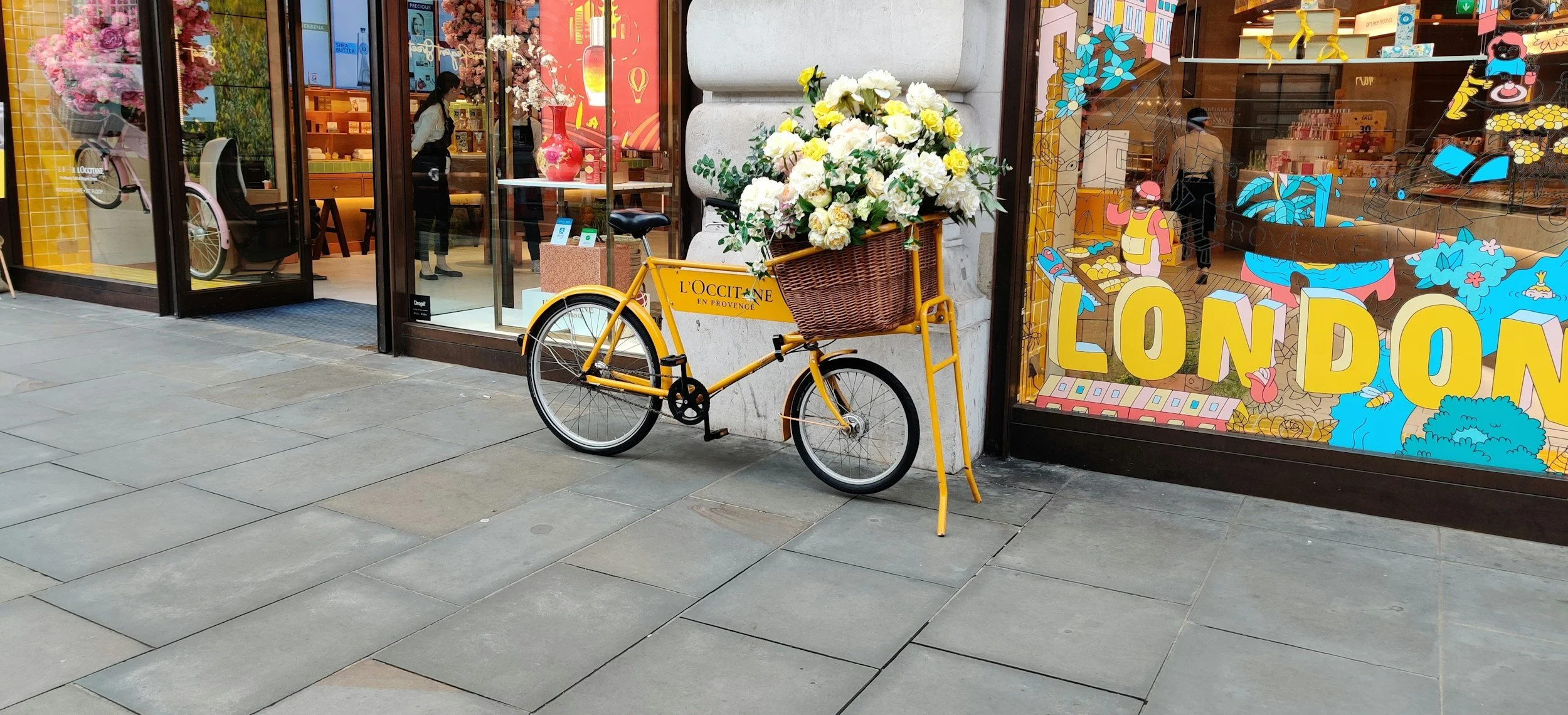 A yellow bicycle with a front basket filled with white and yellow flowers is parked on the sidewalk outside a colorful shop window. The window has bright yellow letters spelling 'LONDON' and various illustrated decorations, with a reflection of the s