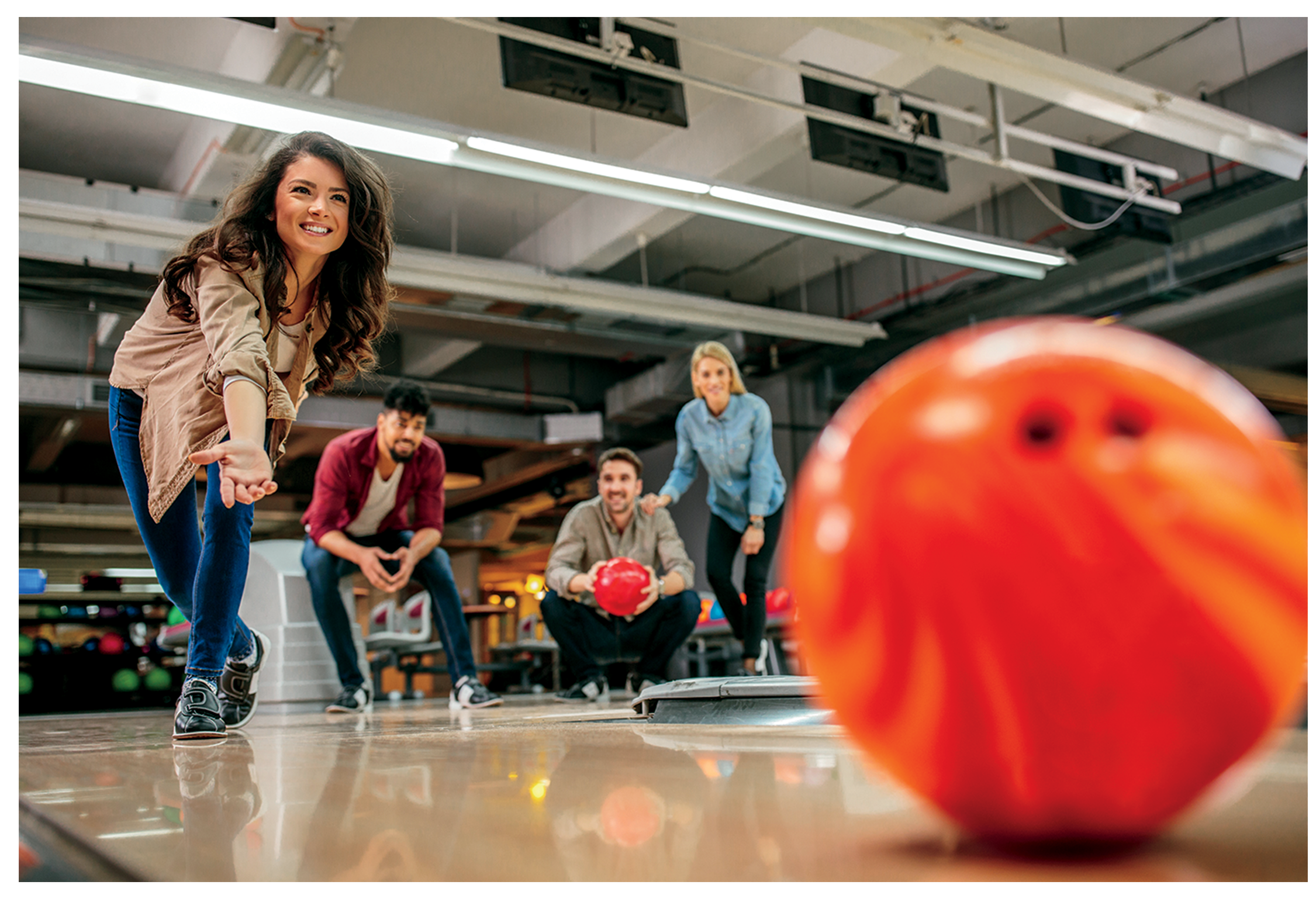 A group of friends bowling indoors, with one woman in focus about to release a bowling ball toward the pins, and three others watching and cheering.