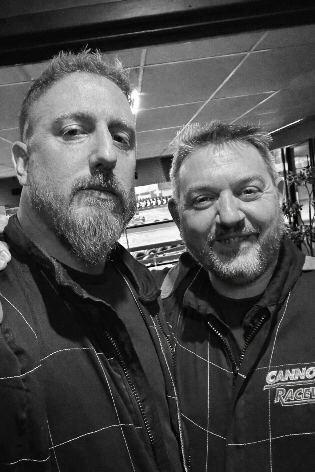Two men with beards smiling, taking a selfie together in a room with a TV and plants in the background.