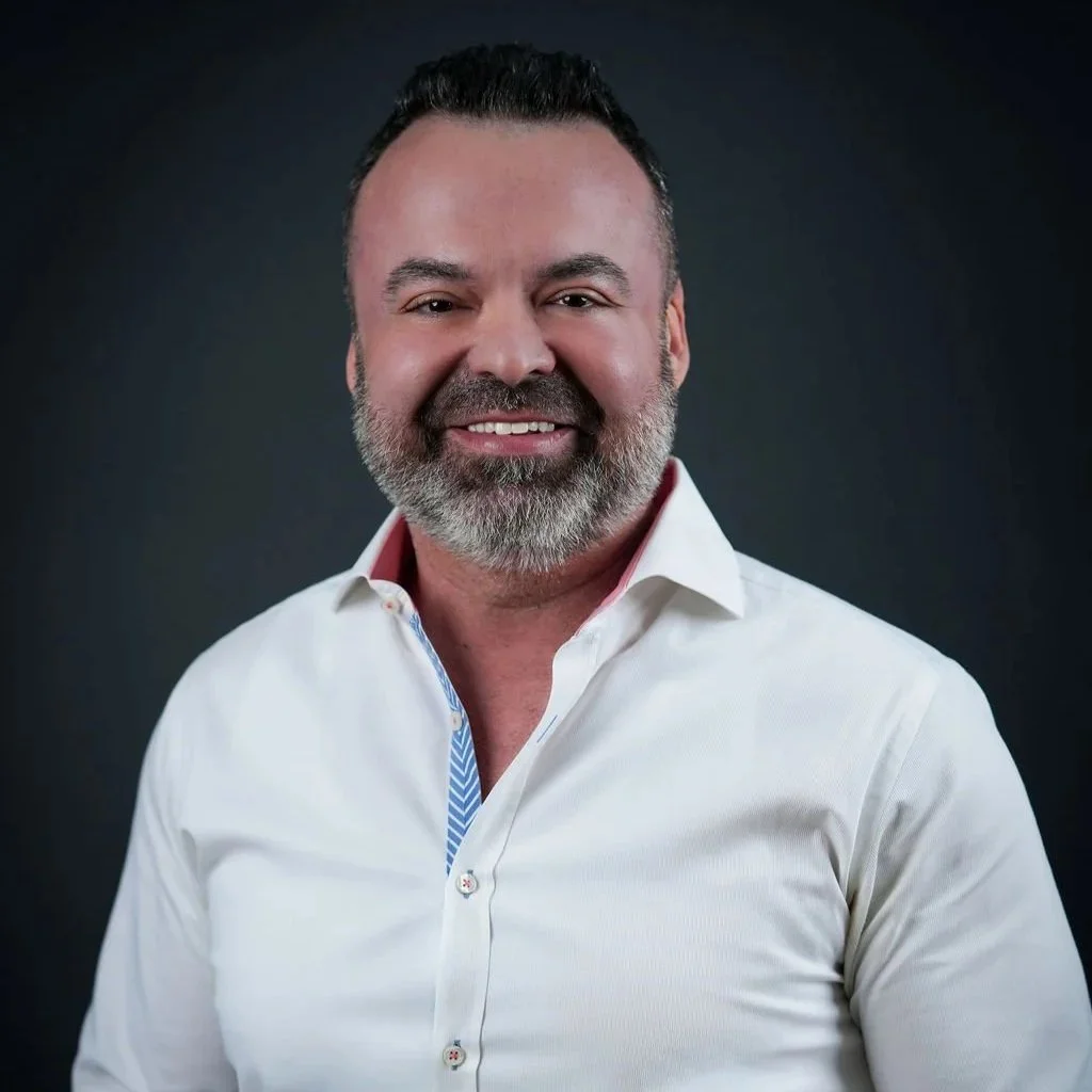 A smiling man with a beard and mustache wearing a white dress shirt, against a dark background.