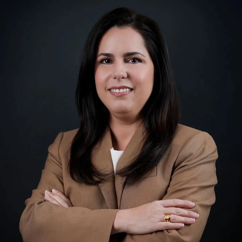 A woman with long dark hair wearing a beige blazer, posing with arms crossed and smiling in front of a dark background.