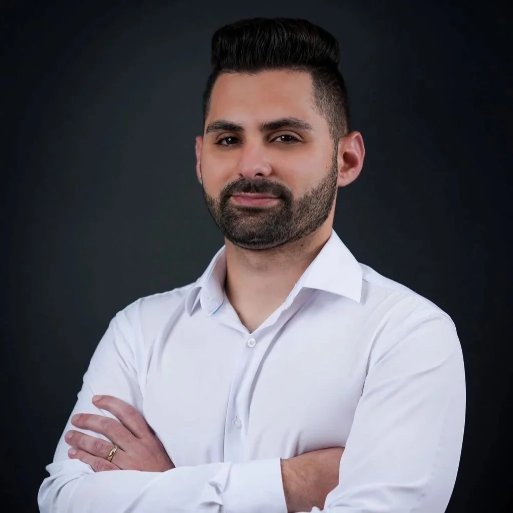 A man with dark hair styled up, a beard, and light skin, wearing a white dress shirt with the top button undone, standing against a dark background with arms crossed.