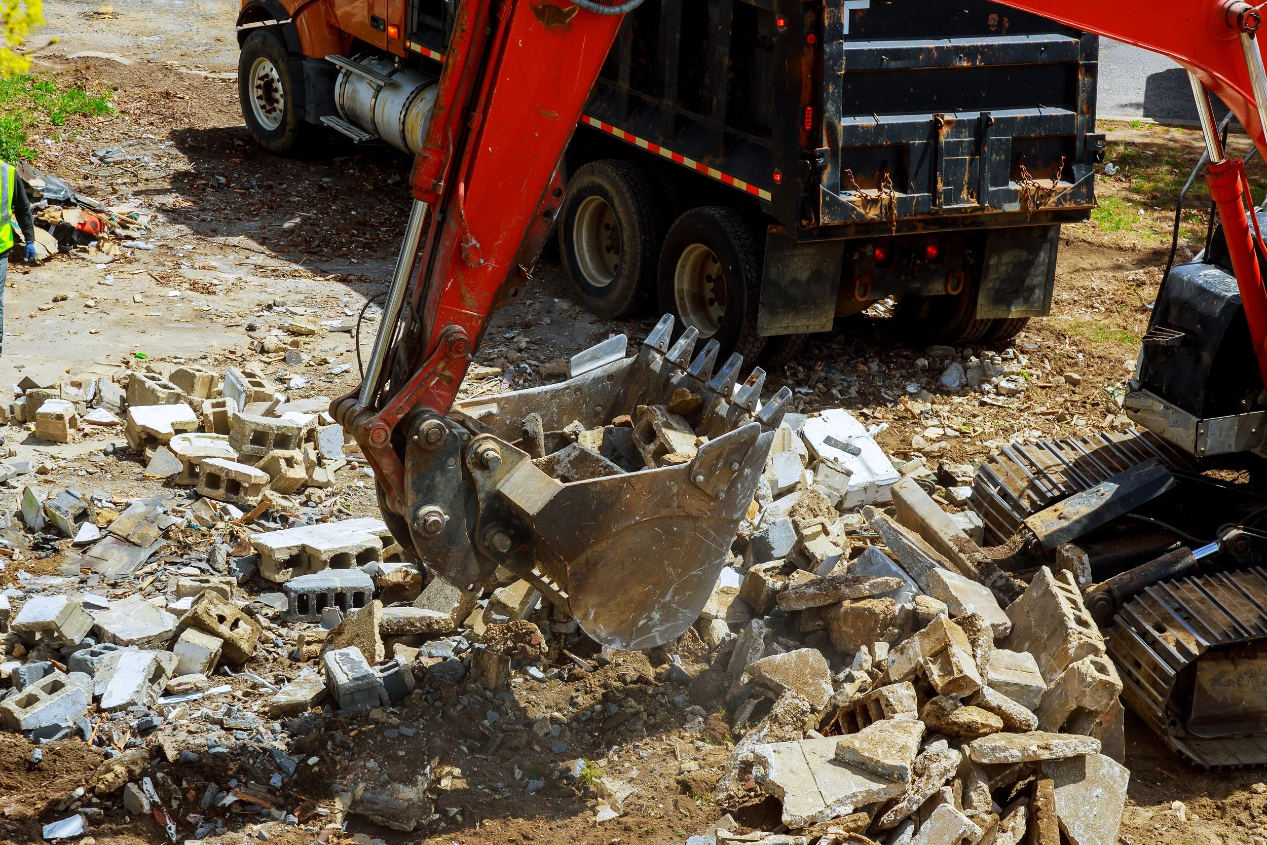 Excavator digging up rubble and debris at a construction site, with a truck in the background.