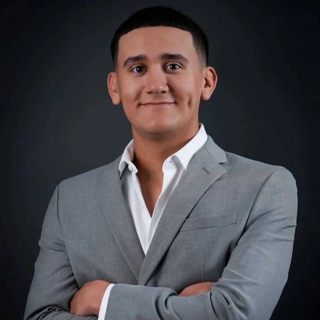 A young man with short dark hair wearing a gray suit and white dress shirt, standing against a dark background, smiling with arms crossed.