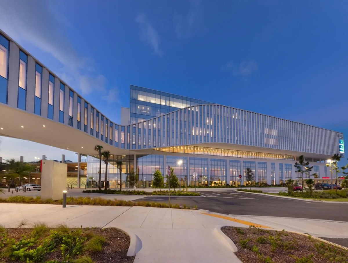 Modern office building with glass exterior and illuminated windows during evening, surrounded by landscaping and parking lot.