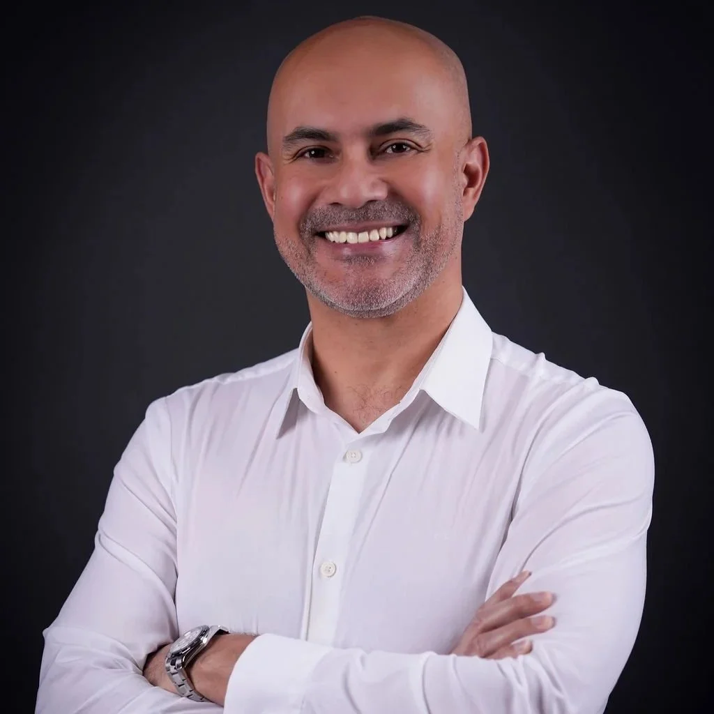 A smiling bald man with a beard in a white shirt standing with arms crossed against a dark background.