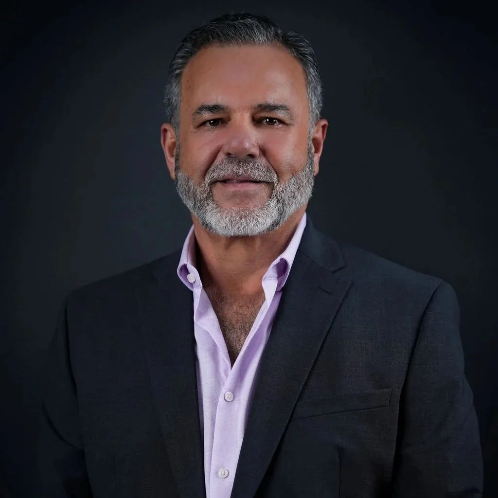 Headshot of a middle-aged man with salt-and-pepper hair and beard, wearing a dark suit jacket and a light purple dress shirt, against a dark background.