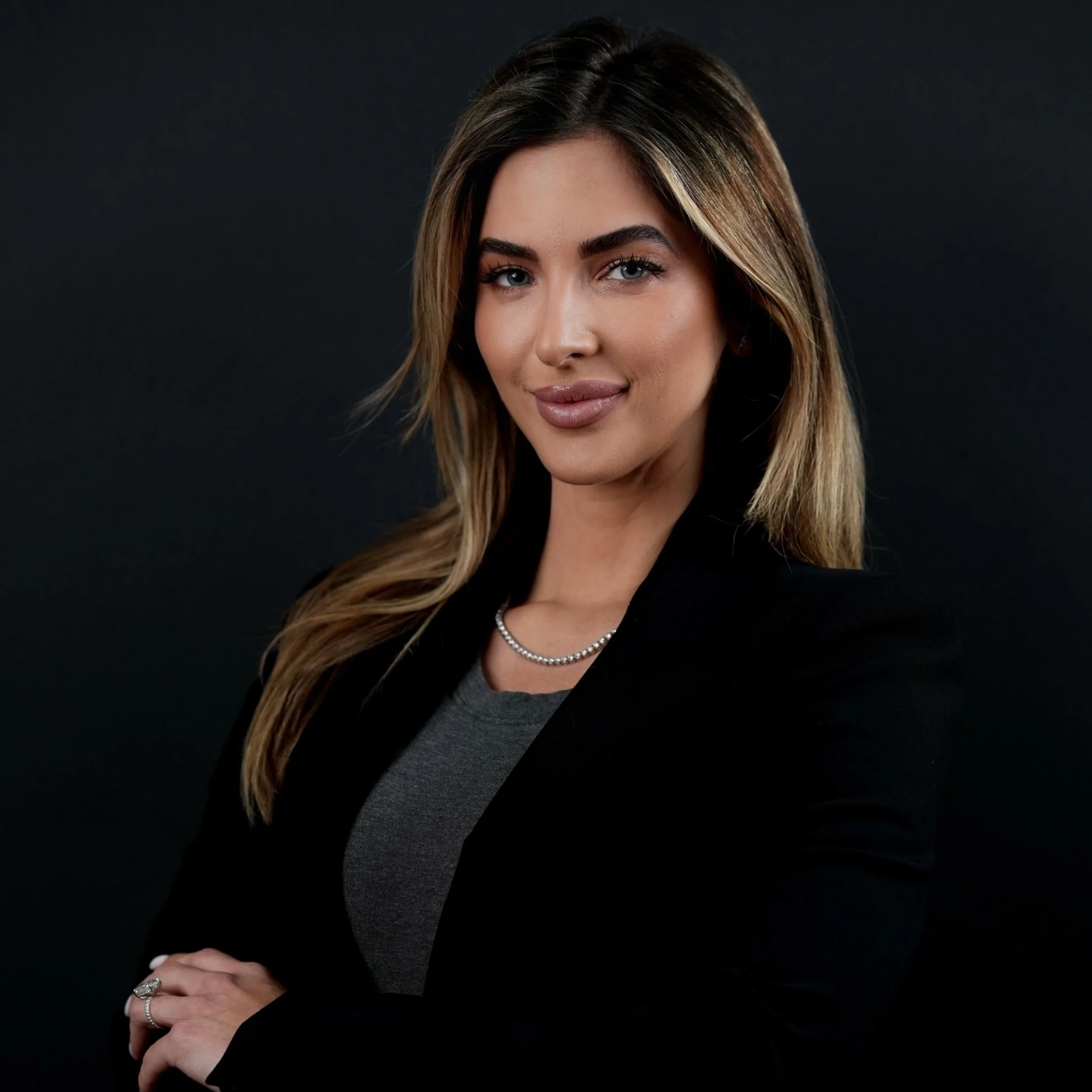 A woman with long, wavy blonde hair, wearing a black blazer over a gray top, accessorized with a pearl necklace and rings, posing against a black background.