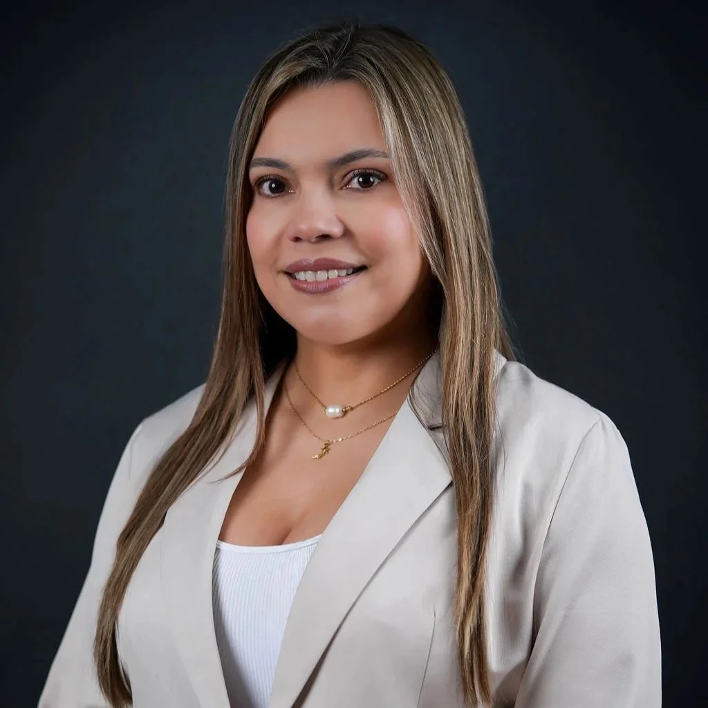 A professional woman with light skin and shoulder-length straight hair, wearing a beige blazer over a white top, and layered necklaces, smiling against a dark background.