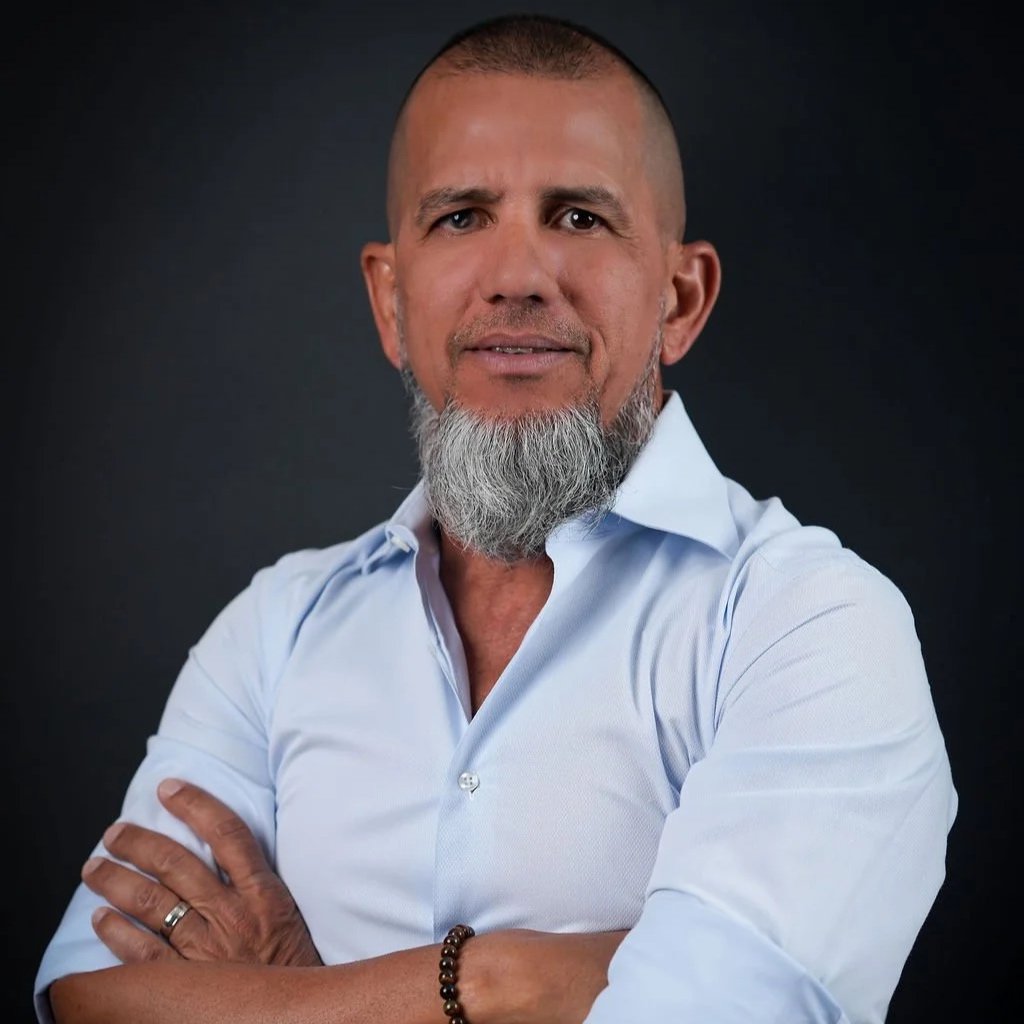 A middle-aged man with a closely shaved head, beard, and mustache, wearing a white button-down shirt with the sleeves rolled up, standing against a dark background with his arms crossed.