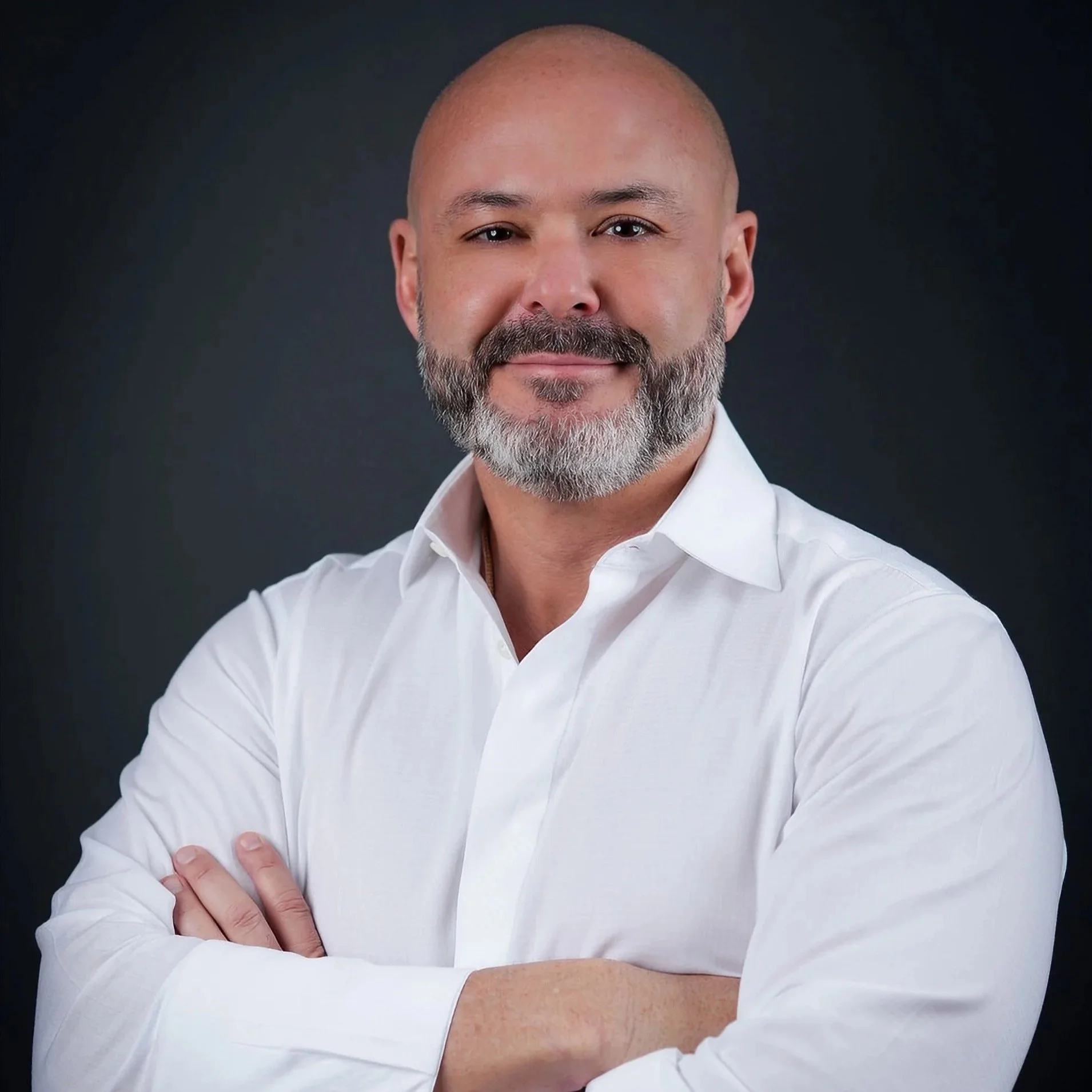 Portrait of a smiling bald man with a beard, wearing a white shirt against a dark background.