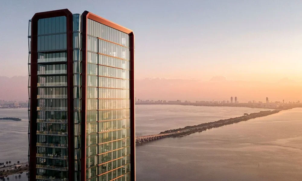A tall modern glass skyscraper with a distinctive red frame, overlooking a body of water during sunset, with a distant city skyline in the background.