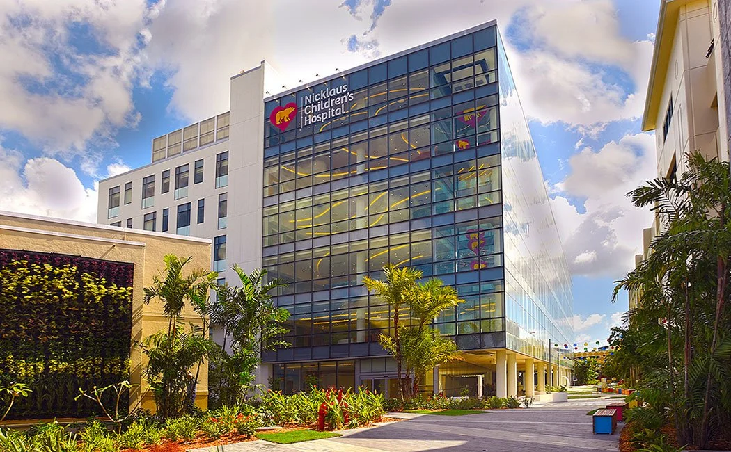Exterior view of Nicklaus Children's Hospital building with glass windows, palm trees, and a cloudy sky.