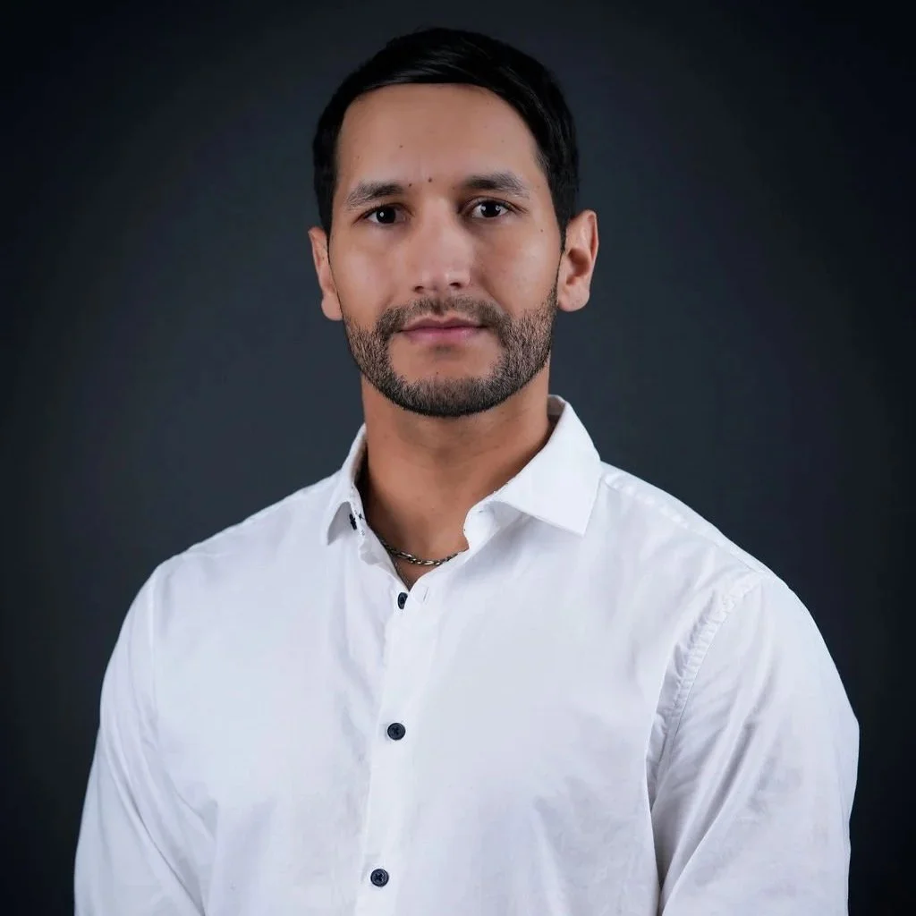 A man with dark hair, a beard, and light skin wearing a white button-up shirt and a chain necklace, standing against a dark background.