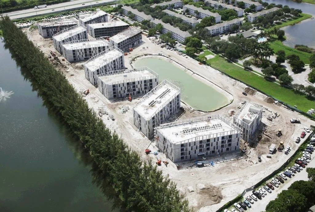 Aerial view of a construction site near a body of water with several modern multi-story buildings in various stages of completion. Green trees and additional buildings are visible in the background.