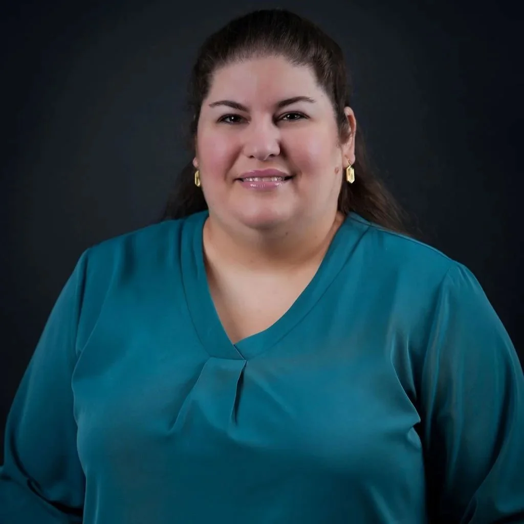 A woman with dark brown hair styled back, wearing a teal blouse and gold earrings, smiling at the camera against a dark background.