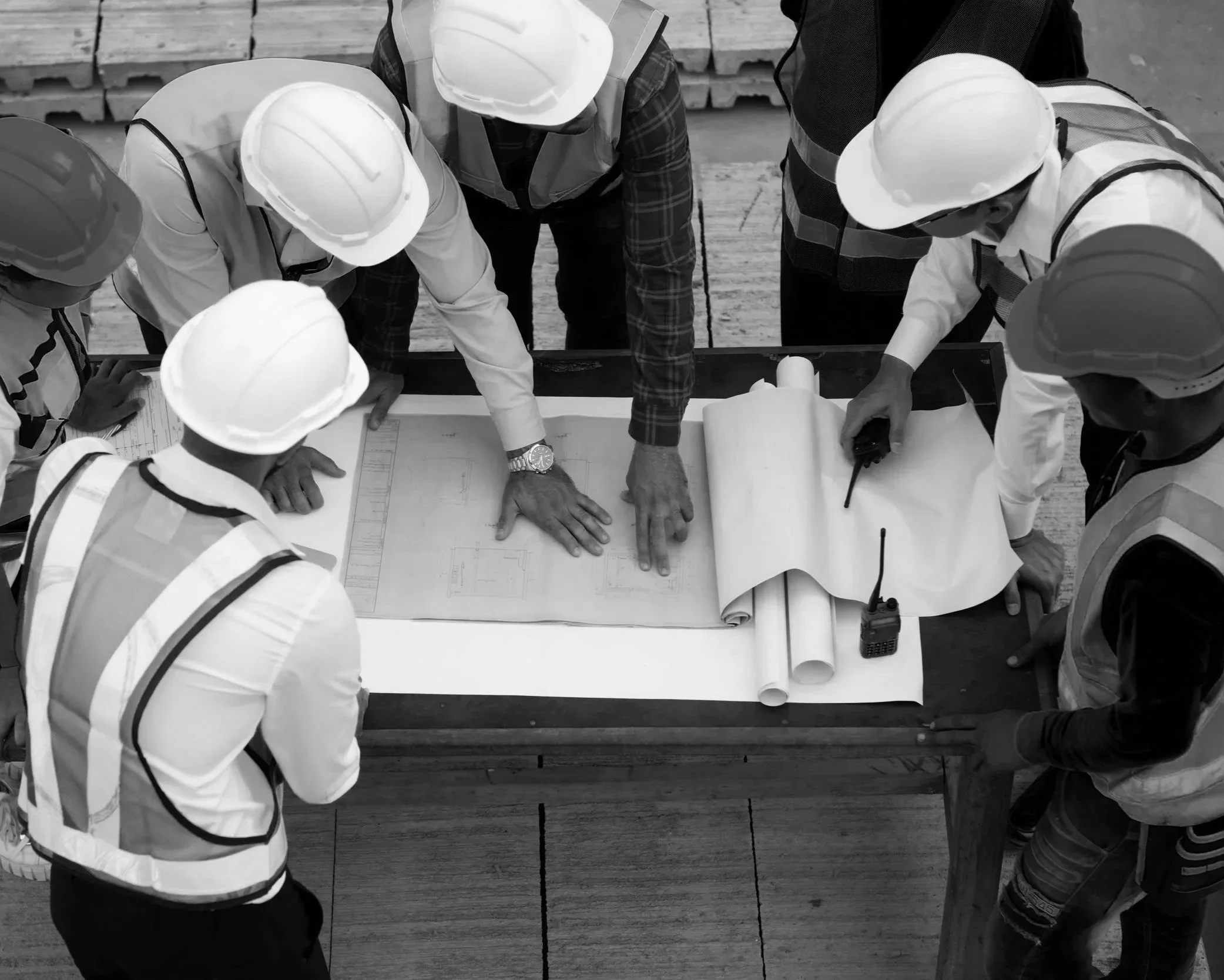 A group of construction workers wearing helmets and safety vests gathered around a table reviewing blueprints, with some pointing and discussing plans, on a construction site.