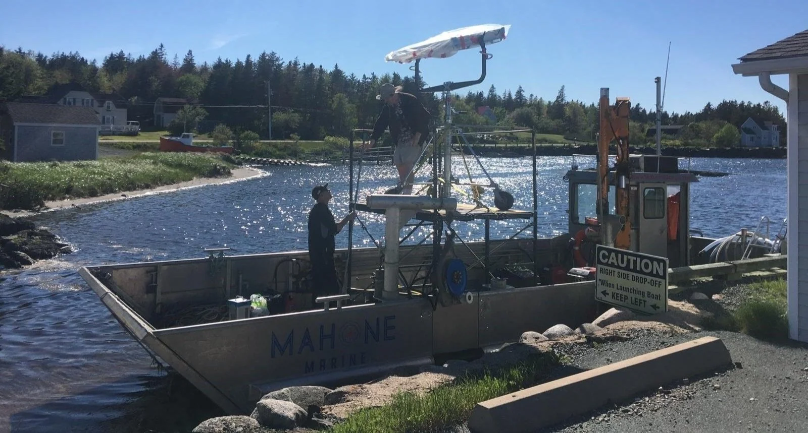 Two people operating a small boat with water testing or monitoring equipment near a shoreline during daytime, with houses and trees in the background.