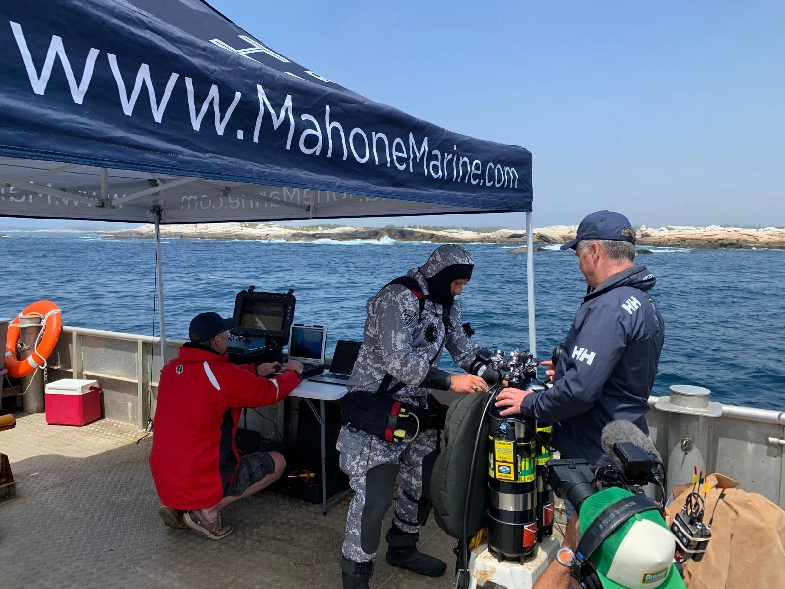 Three people on a boat with equipment, two of them preparing diving gear, one person operating a computer, and a canopy with the website www.Mahonemarine.com on it, along with water and rocky shoreline in the background.