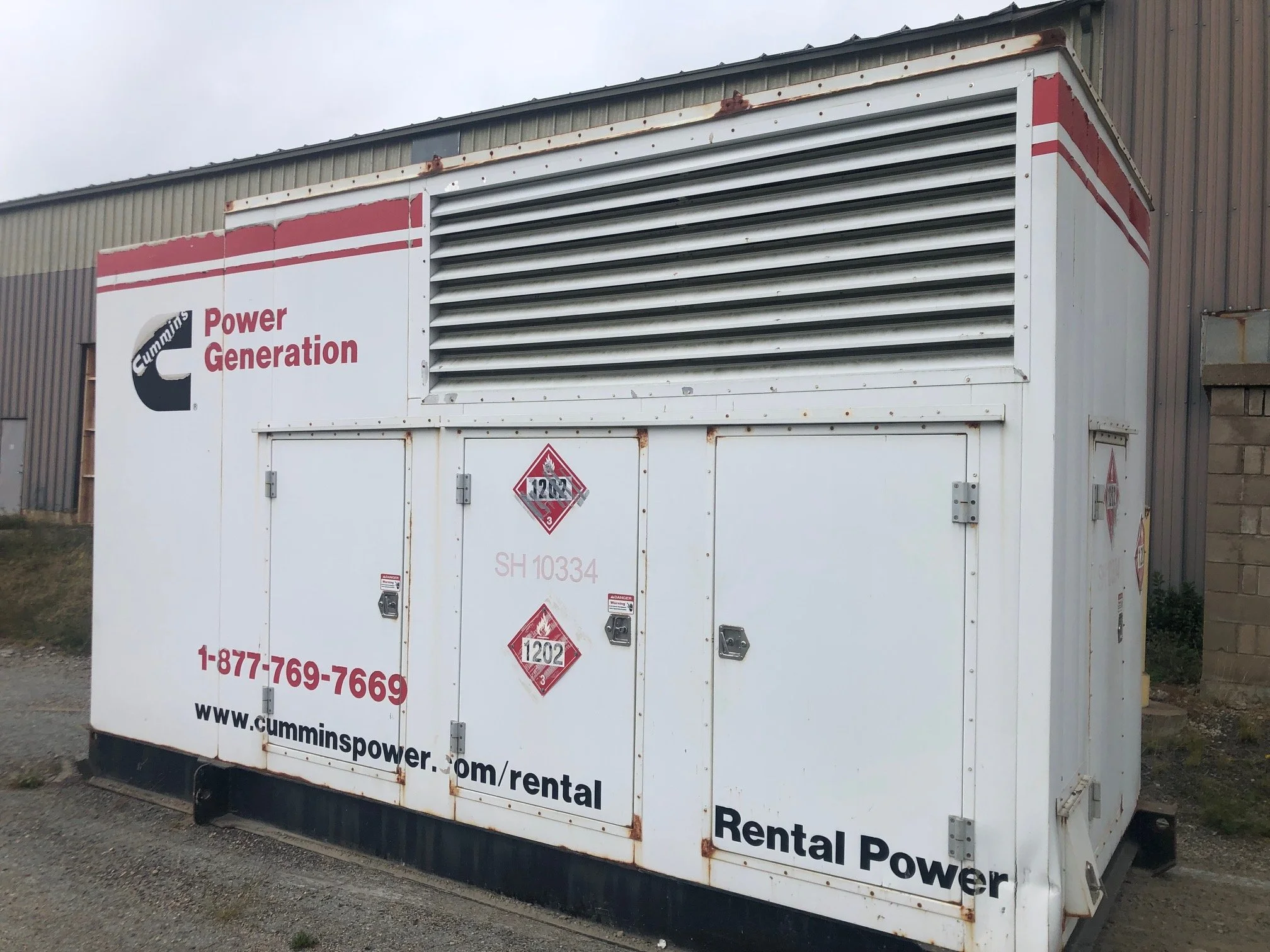 A white commercial generator with red and black signage and labels, including contact information and hazard symbols, situated outdoors next to a metal building.