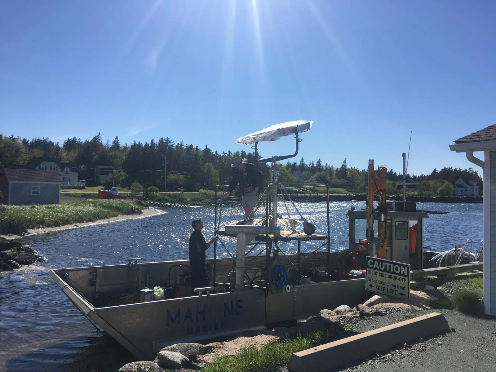 Two workers on a boat named 'Mah One Marine' launching equipment into a river, with houses and trees in the background under a clear, sunny sky.