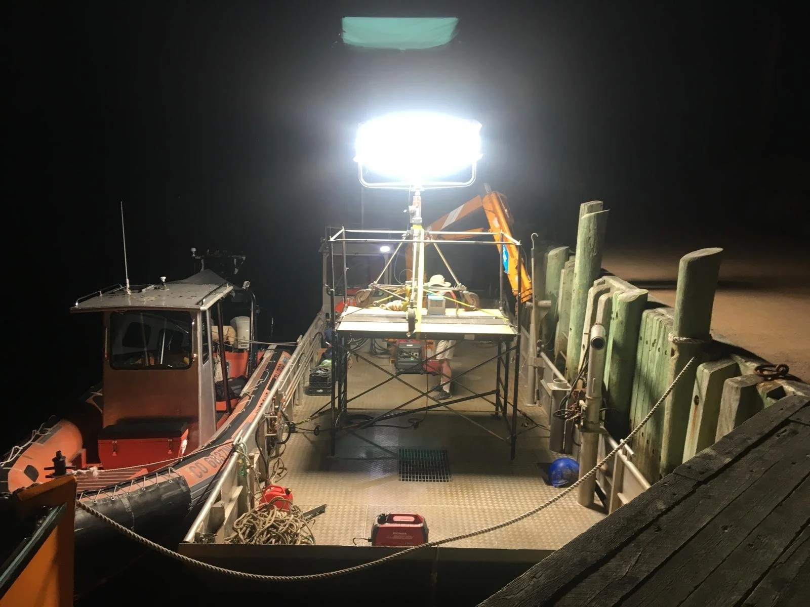 A boat docked at night with a large bright light mounted on a scaffold tower in the center, illuminating the area. The boat is orange with a cabin and various equipment on deck, and there are ropes, chains, and a blue helmet on the deck along with a small red container. The dock has wooden pilings and a wooden platform visible in the foreground.