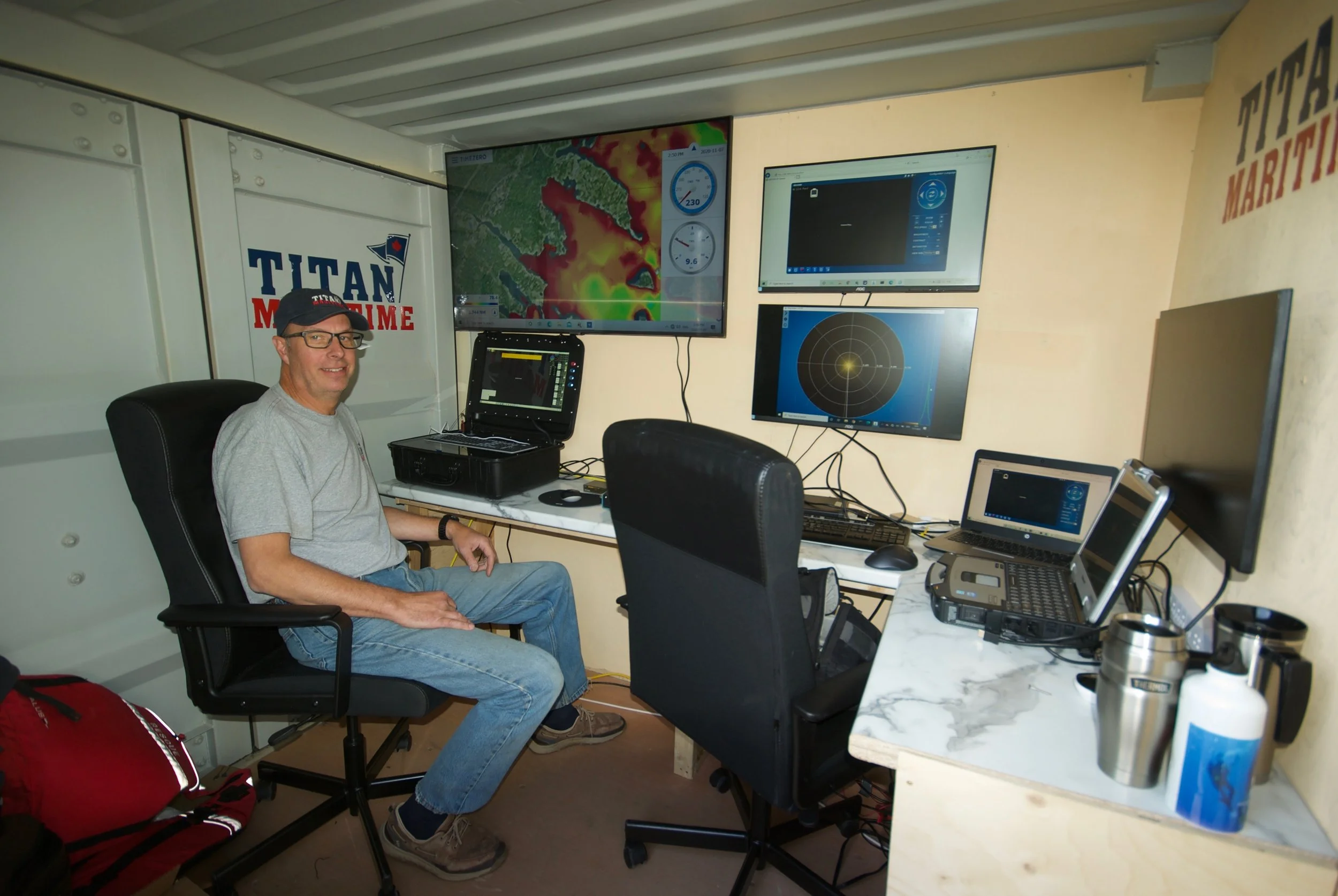 A man sitting in a control room with multiple monitors displaying weather and radar data, wearing glasses, a baseball cap, a gray T-shirt, and jeans.