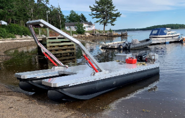A gray and black inflatable boat with a red motor and a metal frame on a shoreline, with another boat docked in the water and trees in the background.