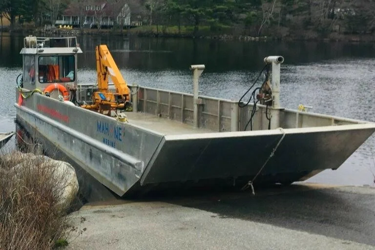 A small flat-bottomed boat with a yellow crane on board, docked near the shore of a river or lake.
