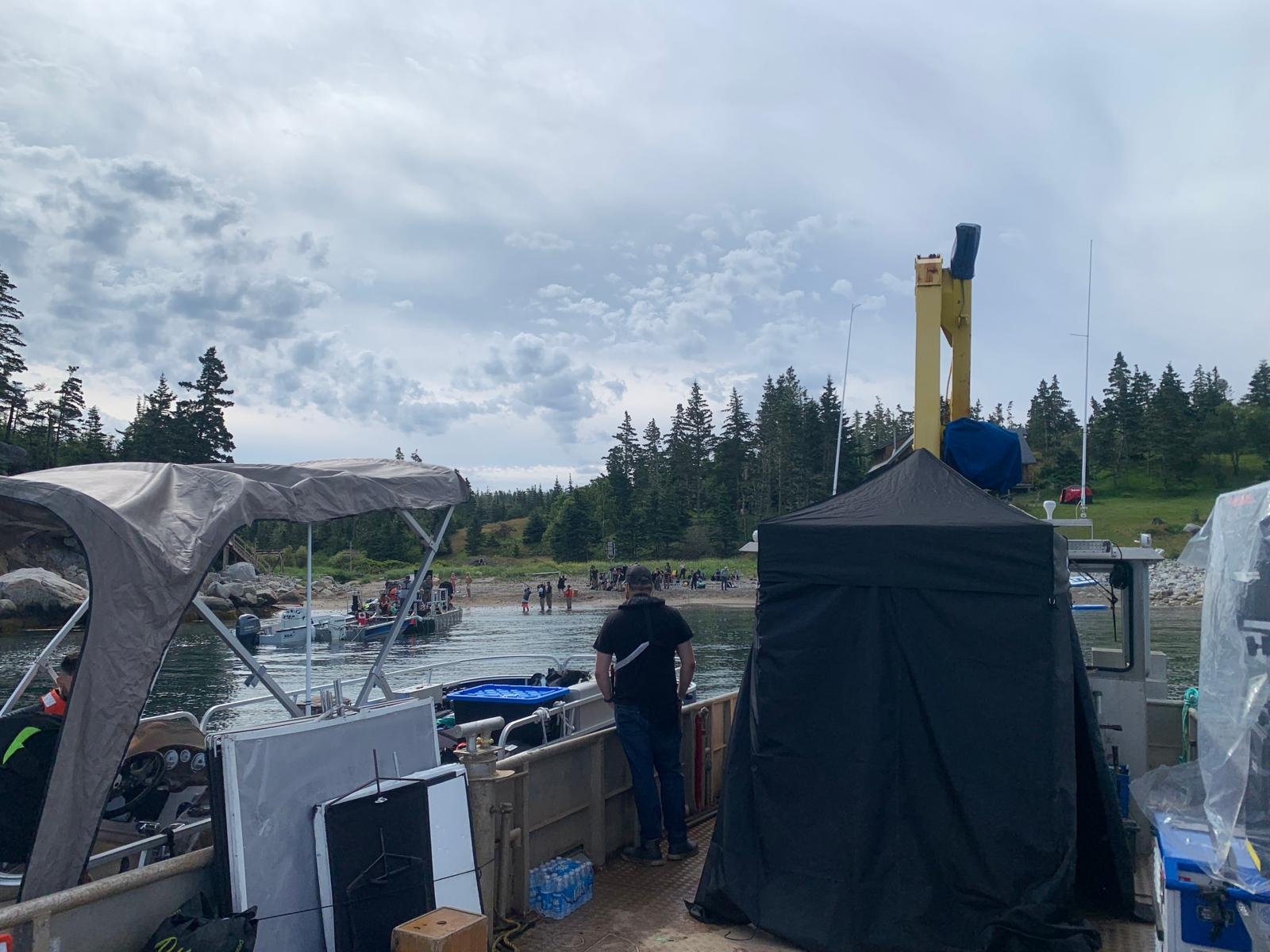 View from a boat showing a dock with other boats, people on a beach, lush green trees on a hillside, and a cloudy sky.