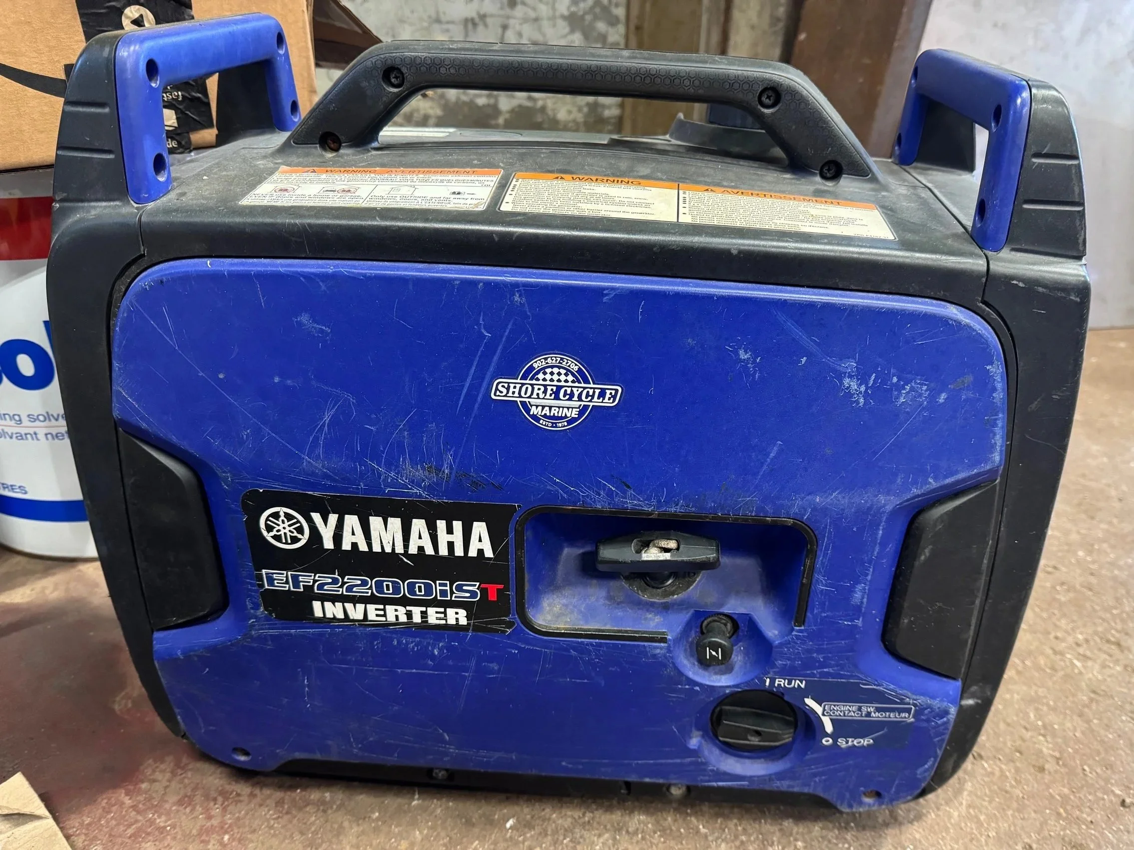Modern wooden house with large glass window in a mountainous landscape, with grass and small bushes in the foreground.