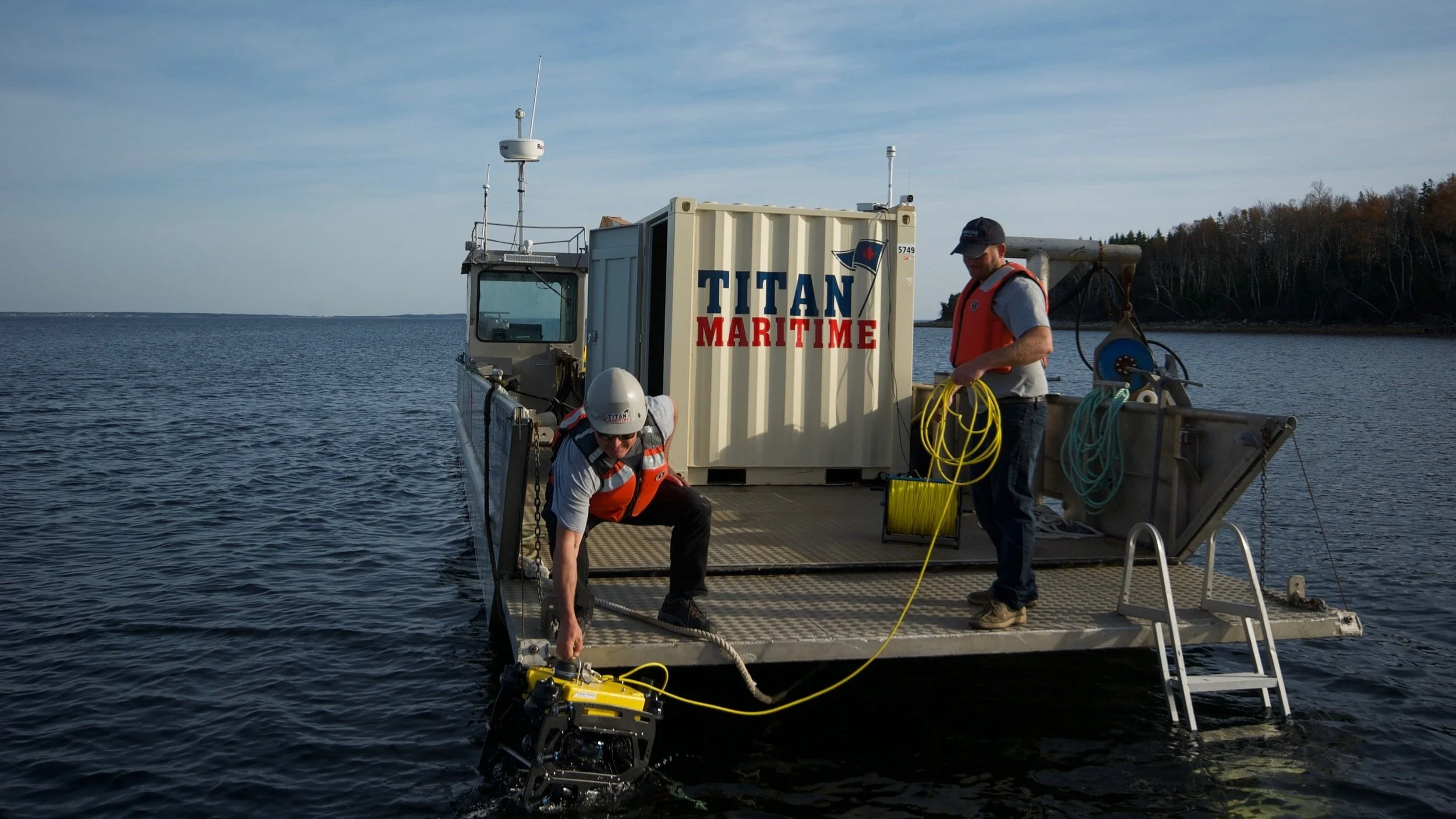 Two workers on a maritime research vessel preparing equipment for deployment in the water, with a body of water and trees in the background.