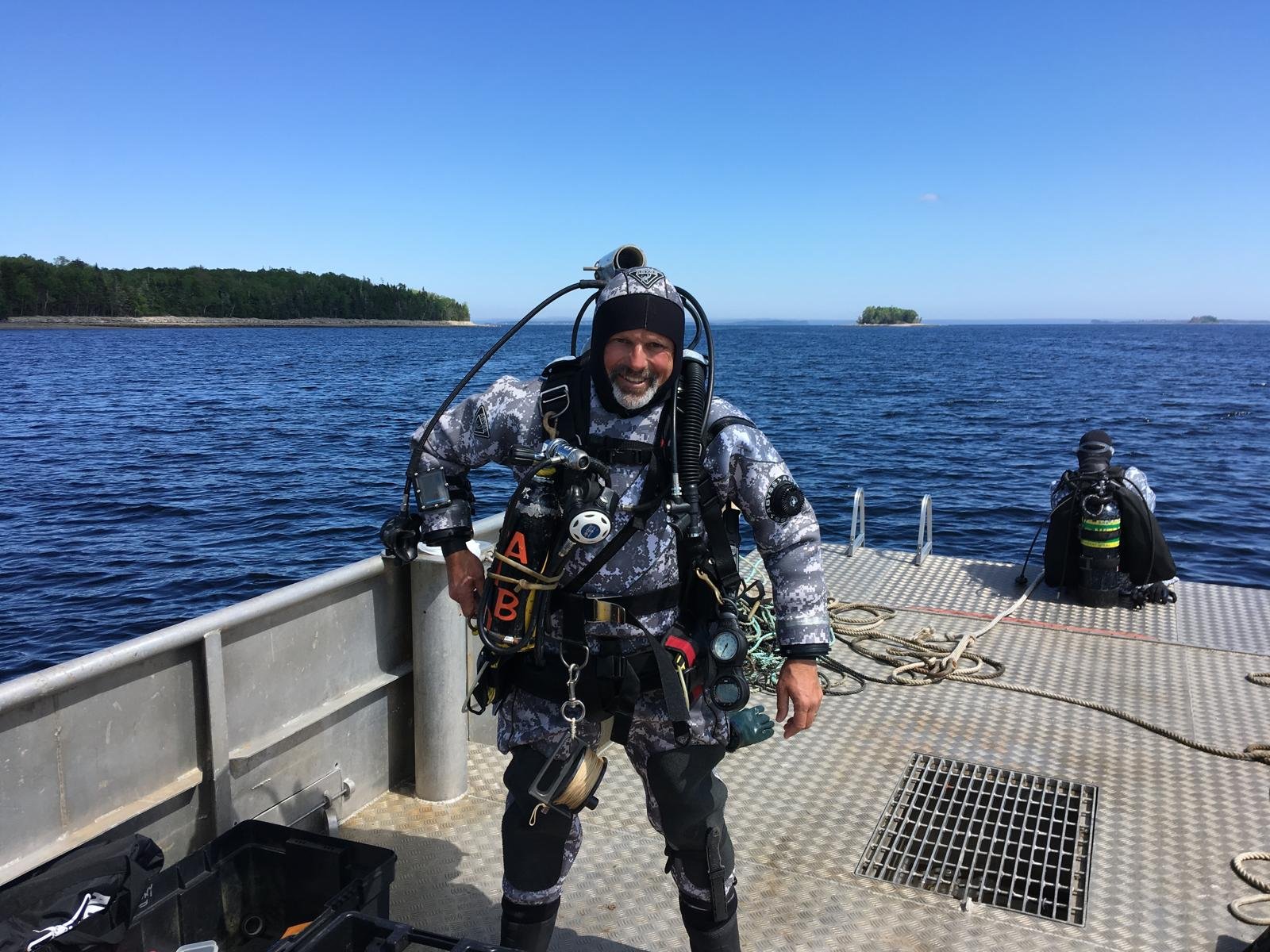 A scuba diver in camouflage gear smiling on a boat with the ocean and islands in the background.
