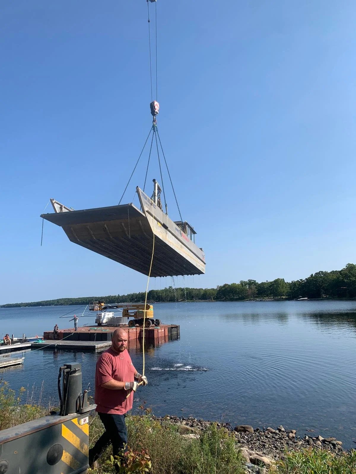 A large boat being lifted out of the water by a crane, with a man standing on the shore holding a rope and wearing gloves.