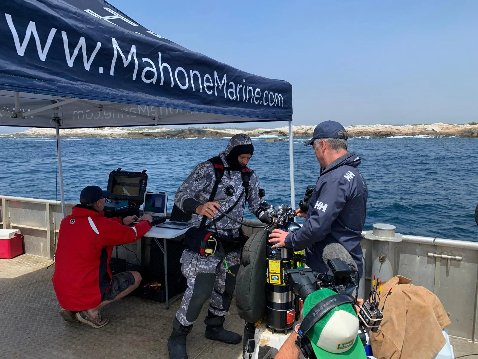 Three men on a boat with diving equipment, one in a camouflage wetsuit preparing gear, another in a navy jacket holding equipment, and a third in a red jacket working on a laptop, with a blue canopy and water in the background.