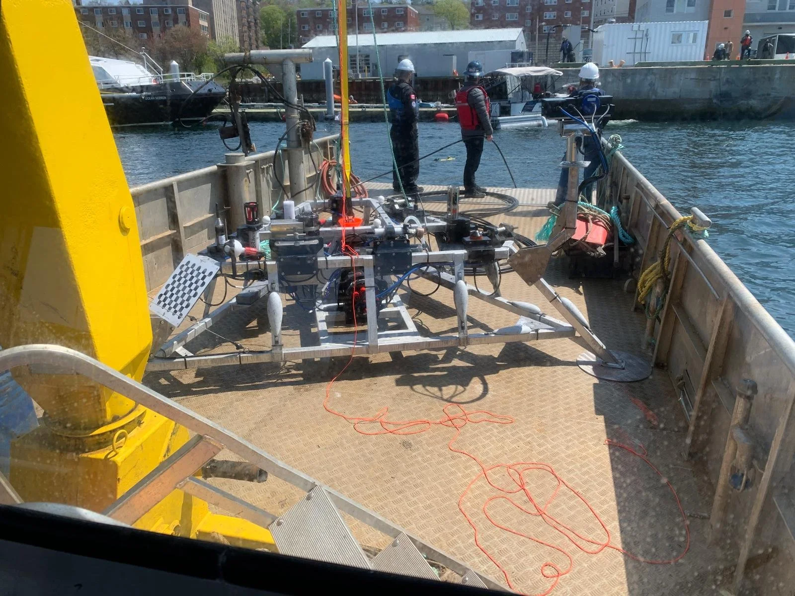 Research equipment and three personnel on a boat, with a cityscape and dock in the background.