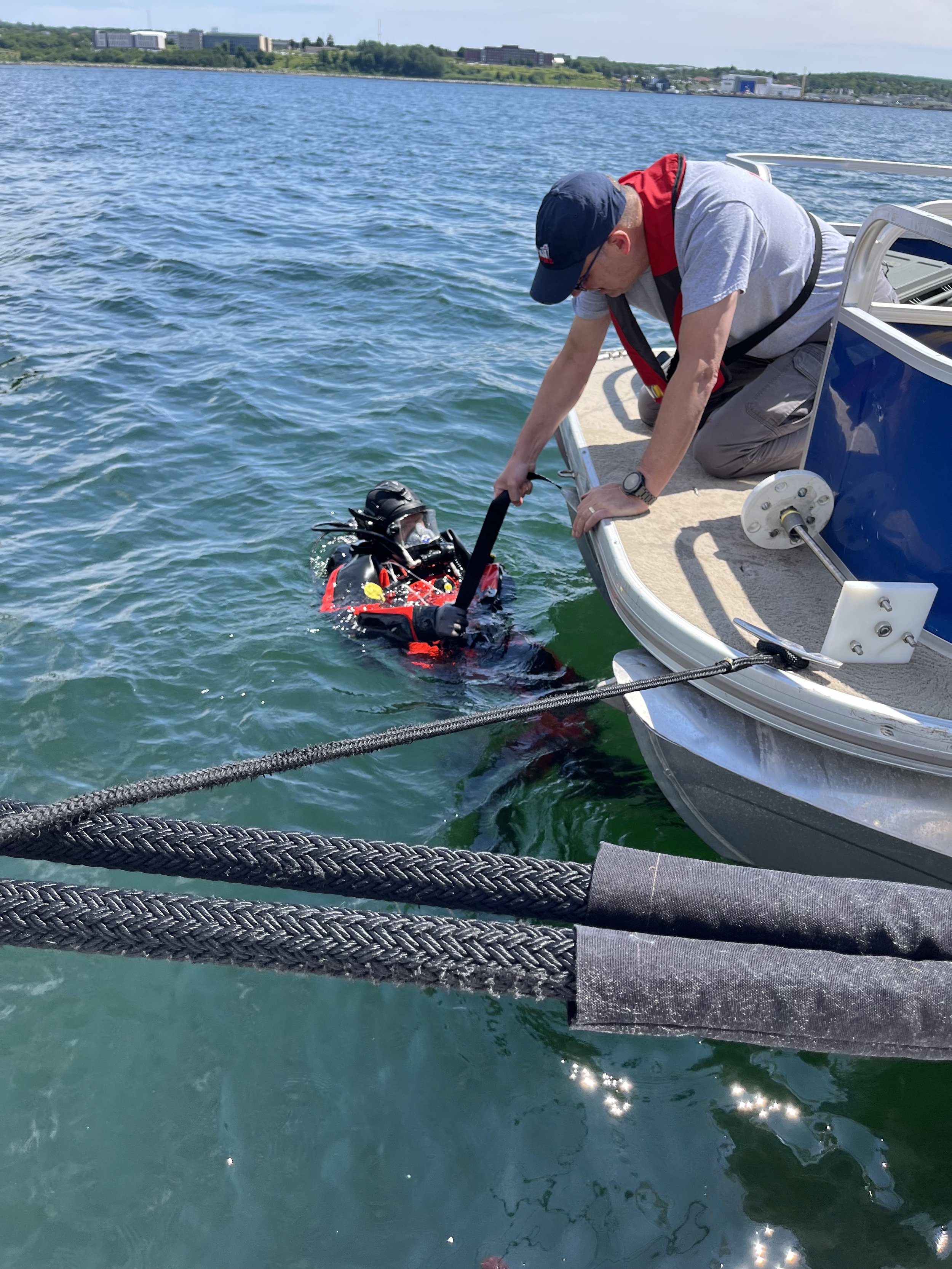 A man in a gray t-shirt, gray pants, and a black cap helps a diver in a red and black wetsuit climb out of the water onto a boat by holding onto a strap. The diver is wearing a helmet and mask, and the scene is set on a body of water with a cityscape in the background.